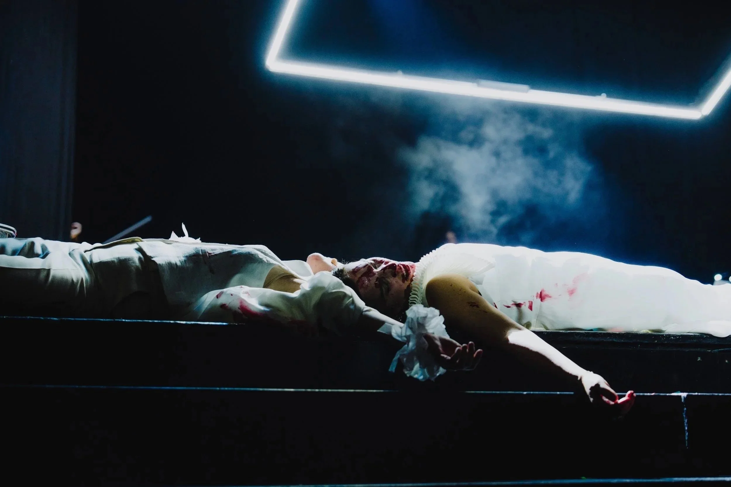 Leiche eines Mannes mit Blutflecken auf einem Bett in einem dunklen Raum, beleuchtet von Neonlicht.