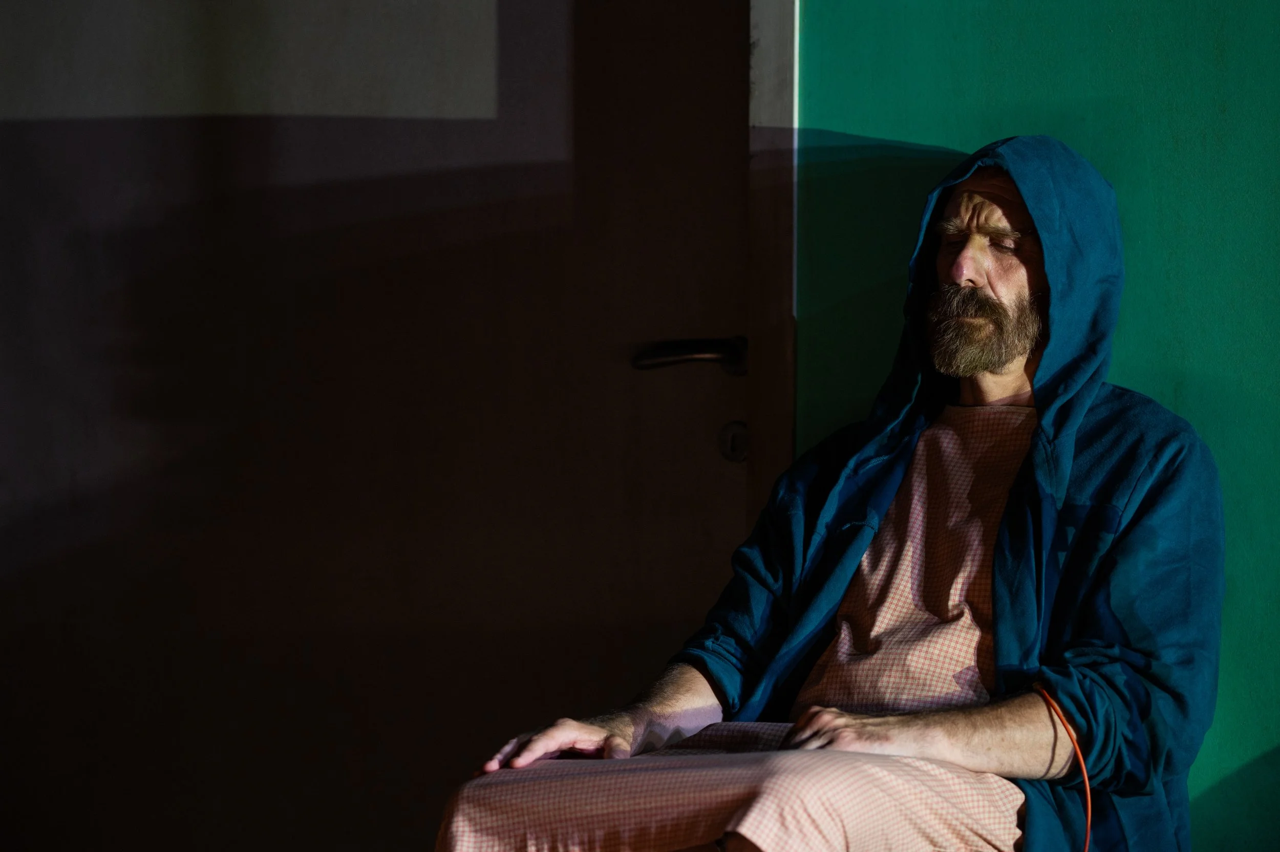 Mann mit Bart, im blauen Hoodie, sitzt vor einer grünen Wand, im Halbdunkel, mit geschlossenen Augen.