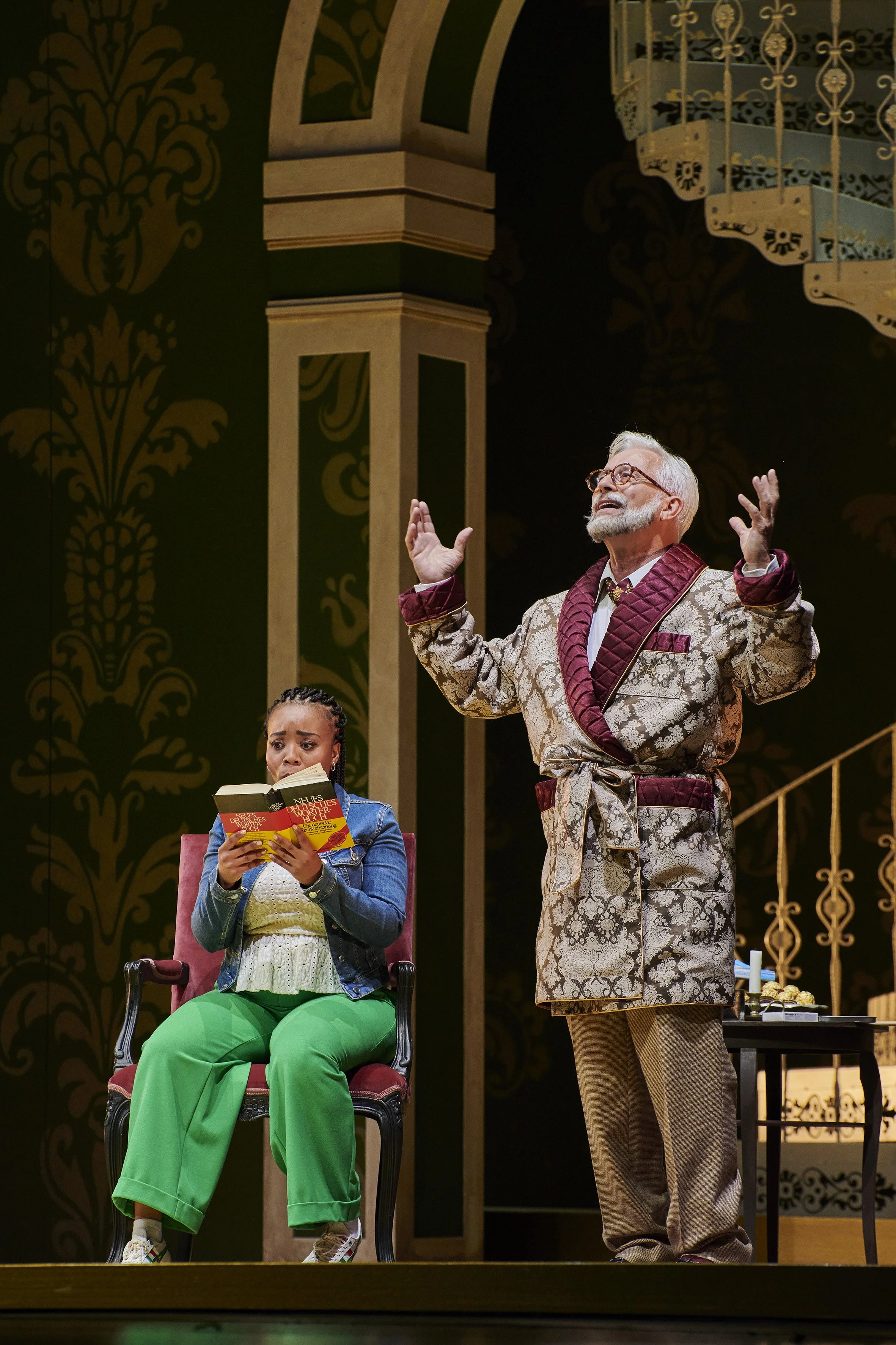 Schauspieler auf Bühne während einer Theateraufführung, eine Frau sitzt in einem roten Sessel, liest ein Buch, ein älterer Mann steht neben ihr mit erhobenen Händen, im Hintergrund goldene und schwarze dekorative Wandverzierungen.
