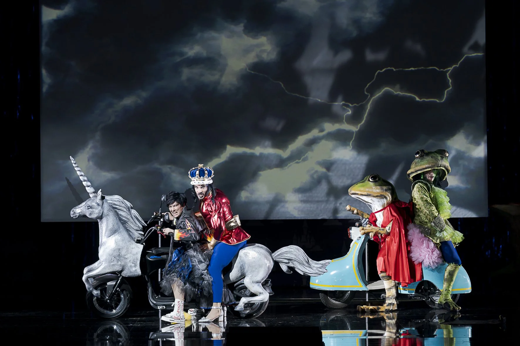 Menschen in Fantasiekostümen mit Tieren und Einhörnern auf einer Bühne, mit Blitz und dunklen Wolken im Hintergrund.