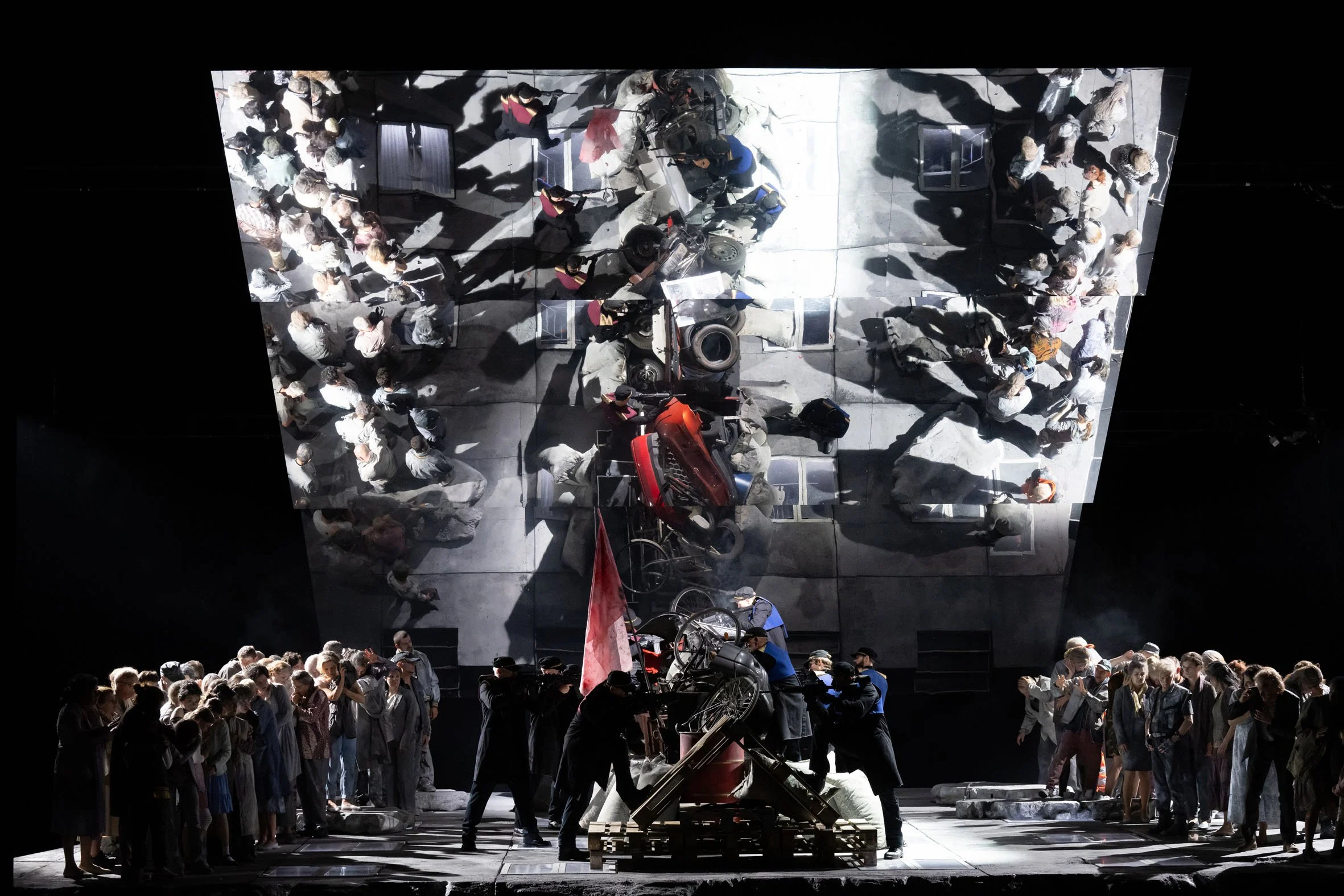 Bühnenbild mit vielen Menschen, einige erscheinen auf einer großen, reflektierenden Wand, andere stehen oder sitzen auf der Bühne im Vordergrund.