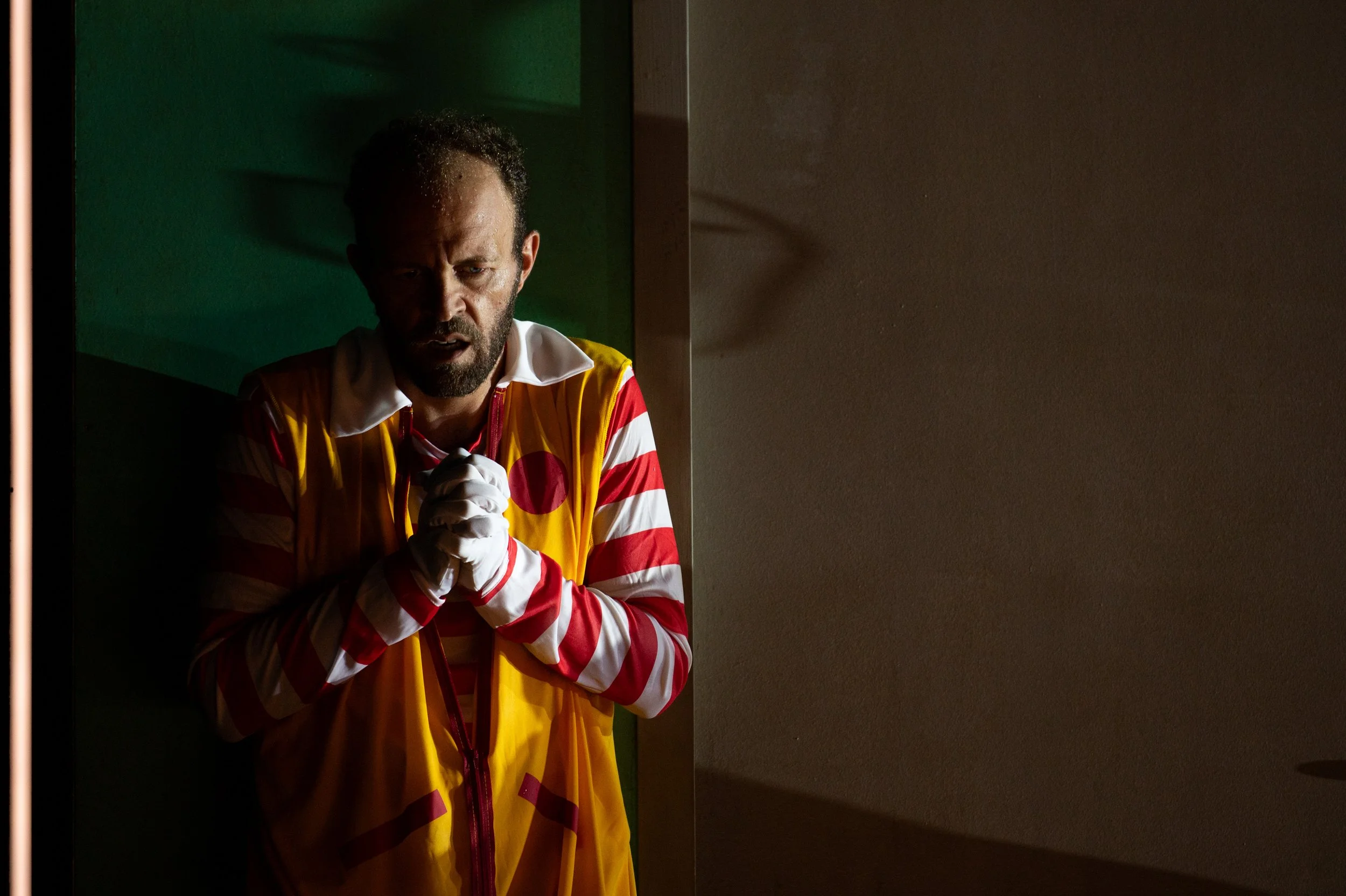 Ein Mann mit dunkler Hautfarbe und Bart steht in einem dunklen Raum, trägt eine bunte, gestreifte Jacke und hat die Hände vor dem Brustkorb verschränkt, in einer nachdenklichen oder besorgten Haltung.