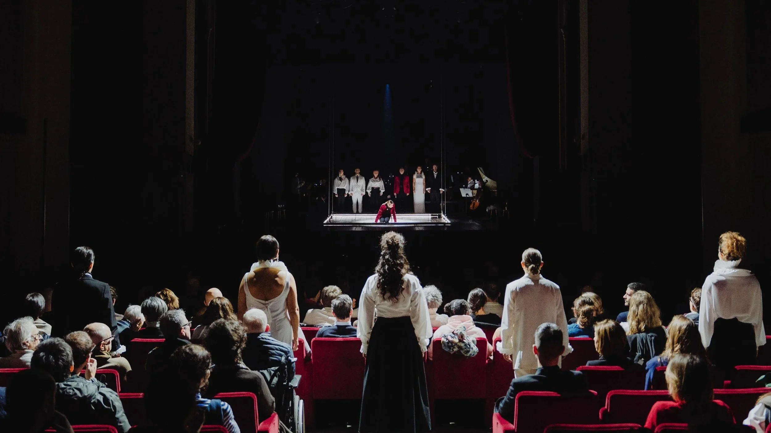 Bühnenauftritt in einem Theater, mit einem Chor auf der Bühne und einer Person, die auf den Knien vor dem Chor sitzt. Das Publikum sitzt in roten Sesseln, einige stehen, die Atmosphäre ist dunkel und konzentriert.
