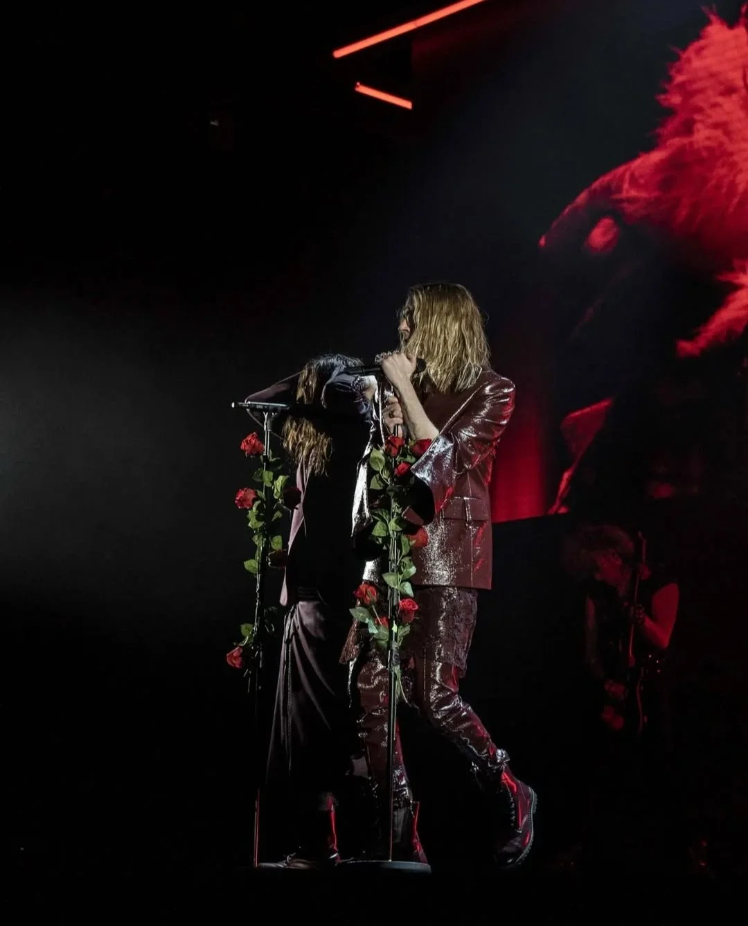 A performer with long blonde hair, wearing a shiny red jacket, sings into a microphone decorated with red roses on a dark stage. Red stage lights are in the background.