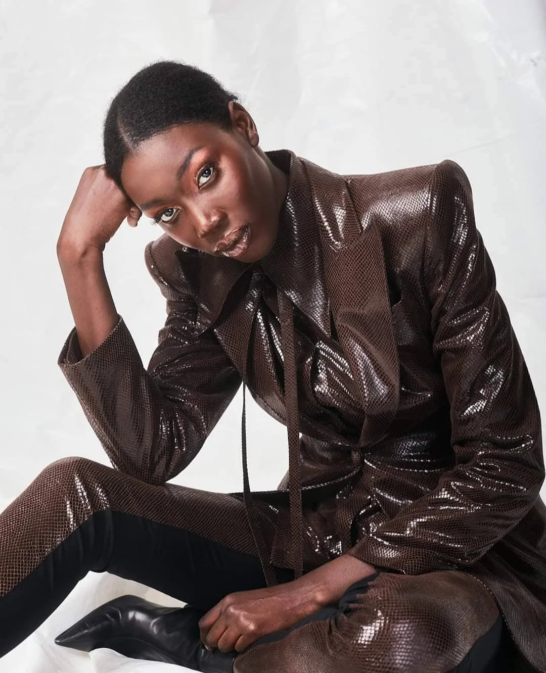 A person with dark skin and short hair posing with one hand resting on their head, wearing a shiny, brown, textured suit and black boots, against a plain white background.