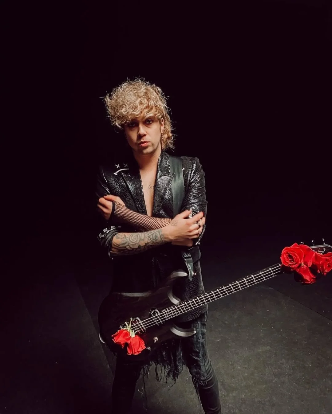 A person with curly blonde hair and tattoos on their arm, wearing a black leather jacket and black ripped jeans, is standing against a black background. They are holding a black guitar decorated with red roses, and they have a serious expression with