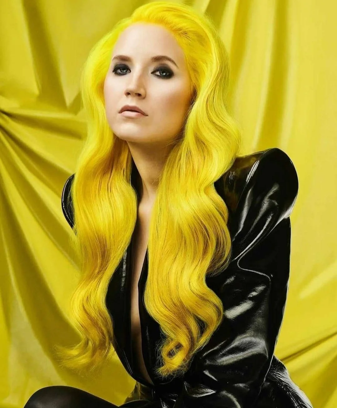 A woman with long, wavy, bright yellow hair poses against a yellow backdrop, wearing a black shiny leather jacket.