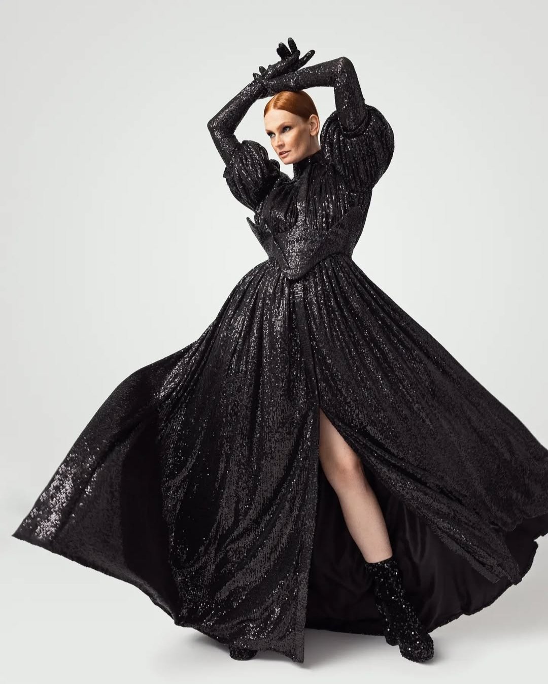 Woman in a black sequined gown with puffed sleeves and a high slit, posing with arms above her head and one leg showing, wearing matching gloves and boots, against a plain white background.