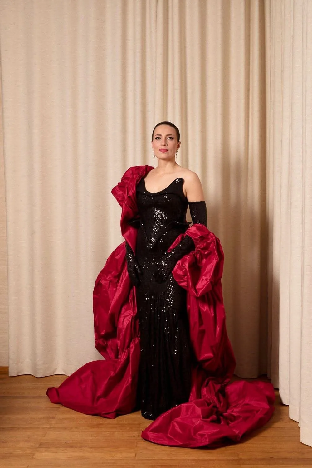 Woman in a black sequin gown with red fabric draped around her, standing in front of beige curtains.