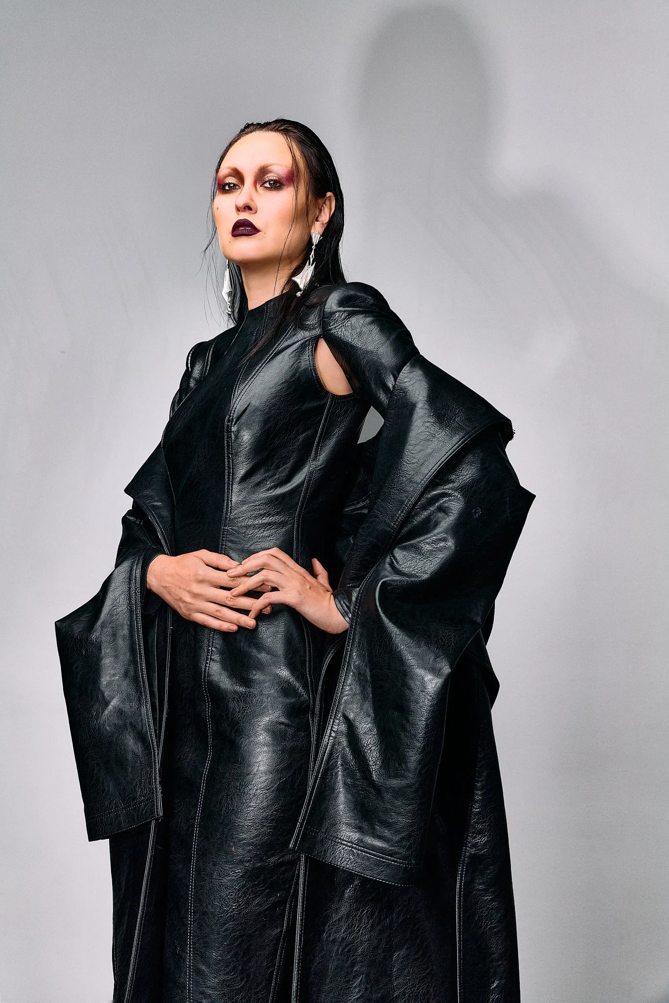 A woman with long black hair, dark makeup, and earrings, wearing a black leather outfit, posing with her hands on her hips against a plain gray background.