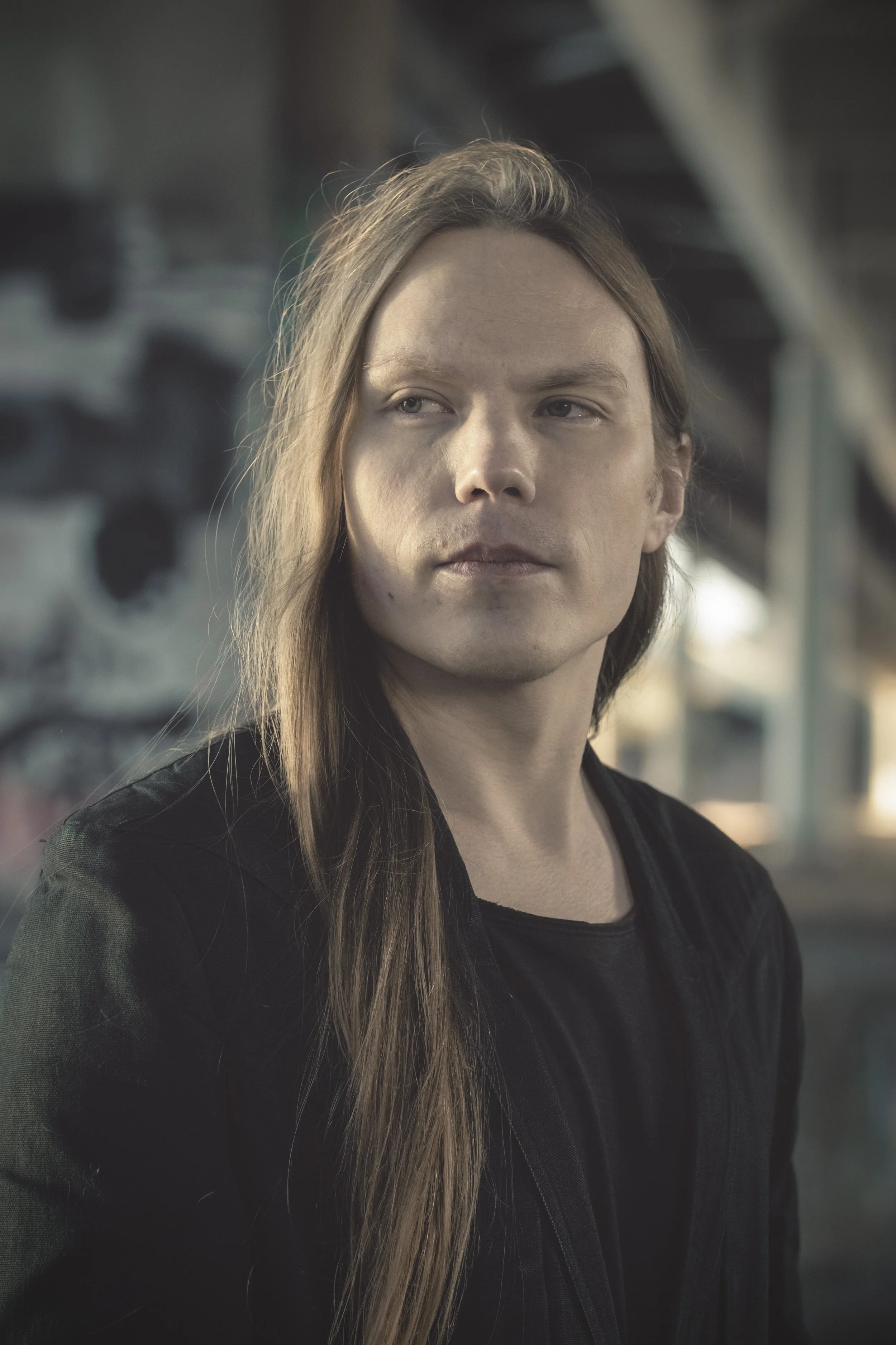 A young man with long brown hair and a pale complexion, wearing a black jacket, stands outdoors with an urban, graffiti-covered background.
