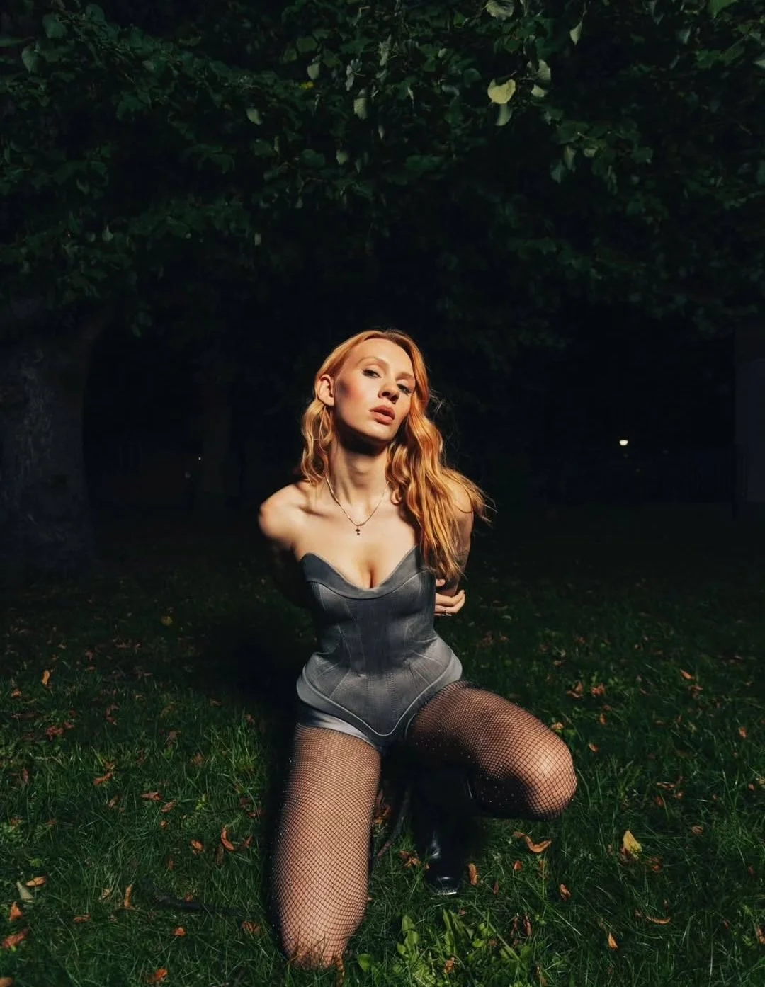 A woman with long red hair, wearing a gray corset and fishnet stockings, kneels on grass under a large tree at night.
