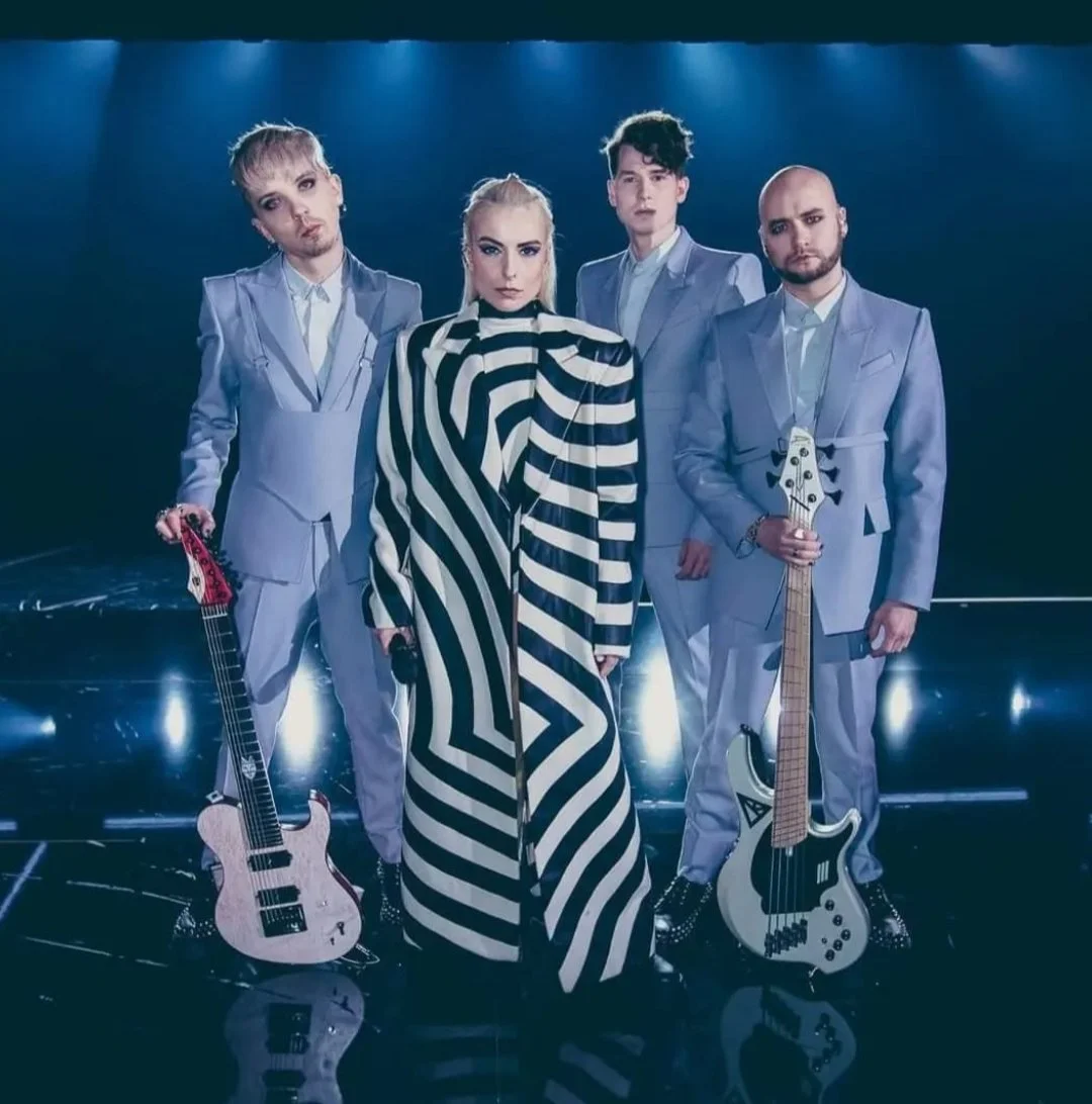 A band of four members posing on stage with musical instruments, including guitars and a bass, under blue stage lights. The members are dressed in coordinated light blue suits, and the woman in the center wears a black-and-white striped outfit.