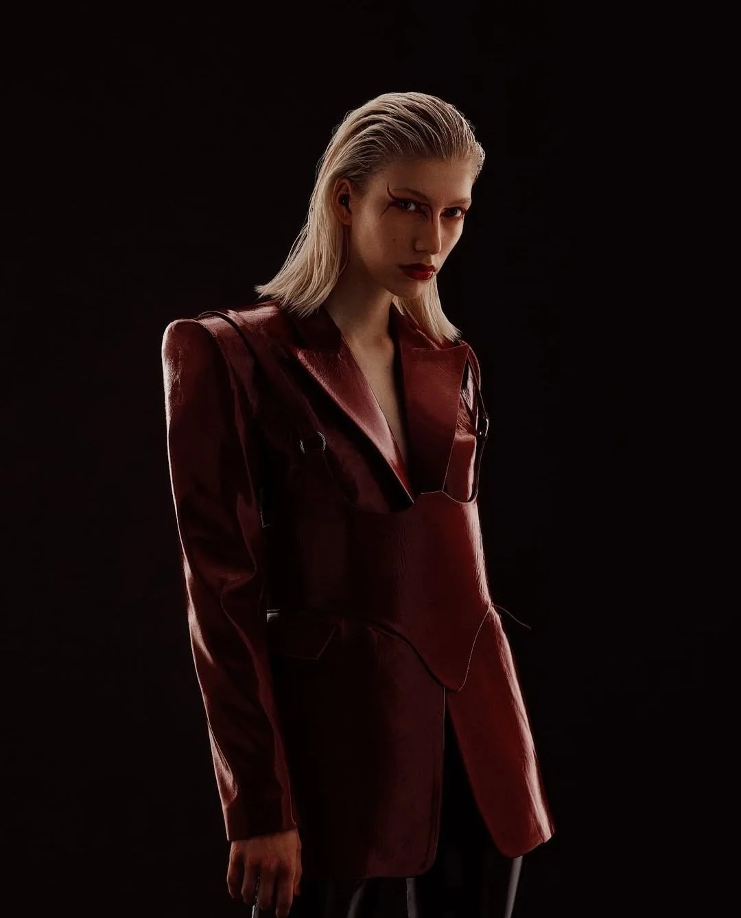 A woman with platinum blonde hair, wearing a dark red leather jacket, poses against a black background with a serious expression.