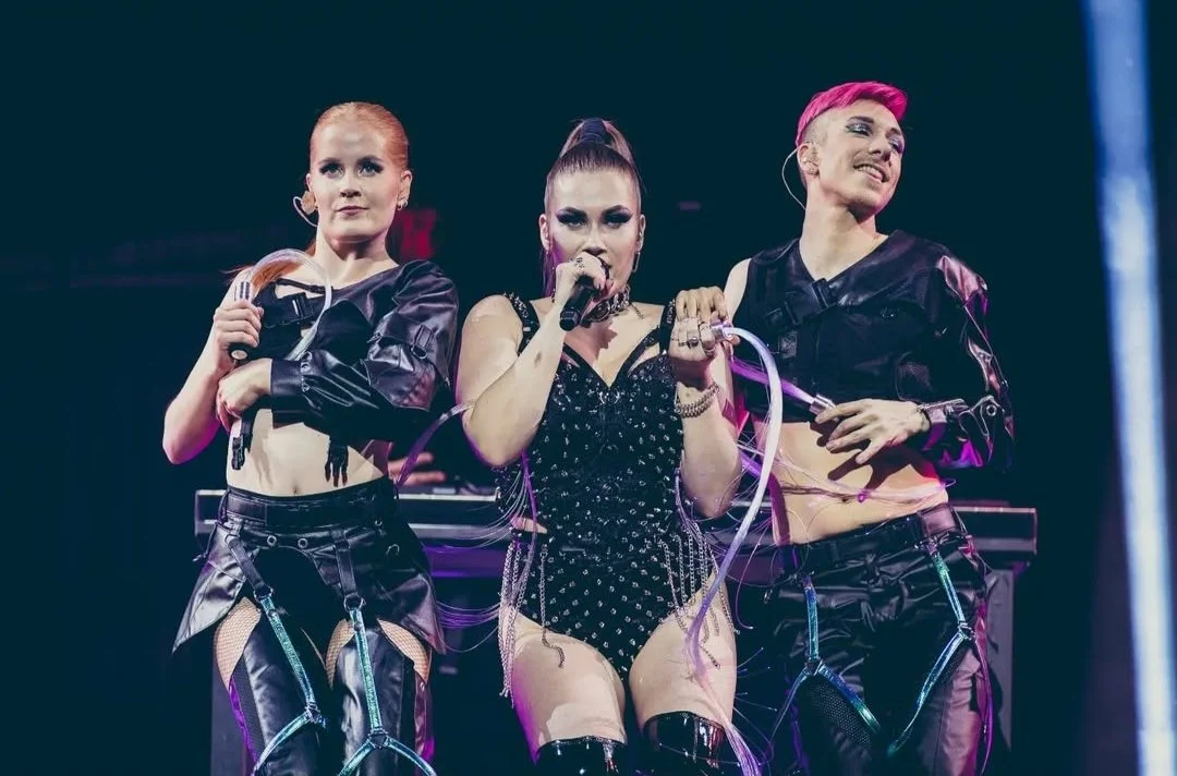 Three female performers on stage, two holding microphones, dressed in black leather outfits with chains and bondage-inspired accessories, one with pink hair and a black dress with silver embellishments, performing with a DJ in the background.