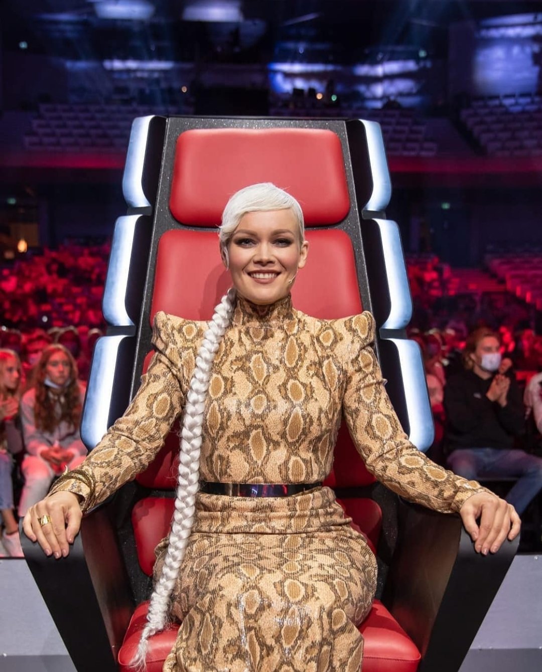 A woman with platinum blonde hair styled in a long braid sitting in a large red and black chair on a stage with an audience in the background.