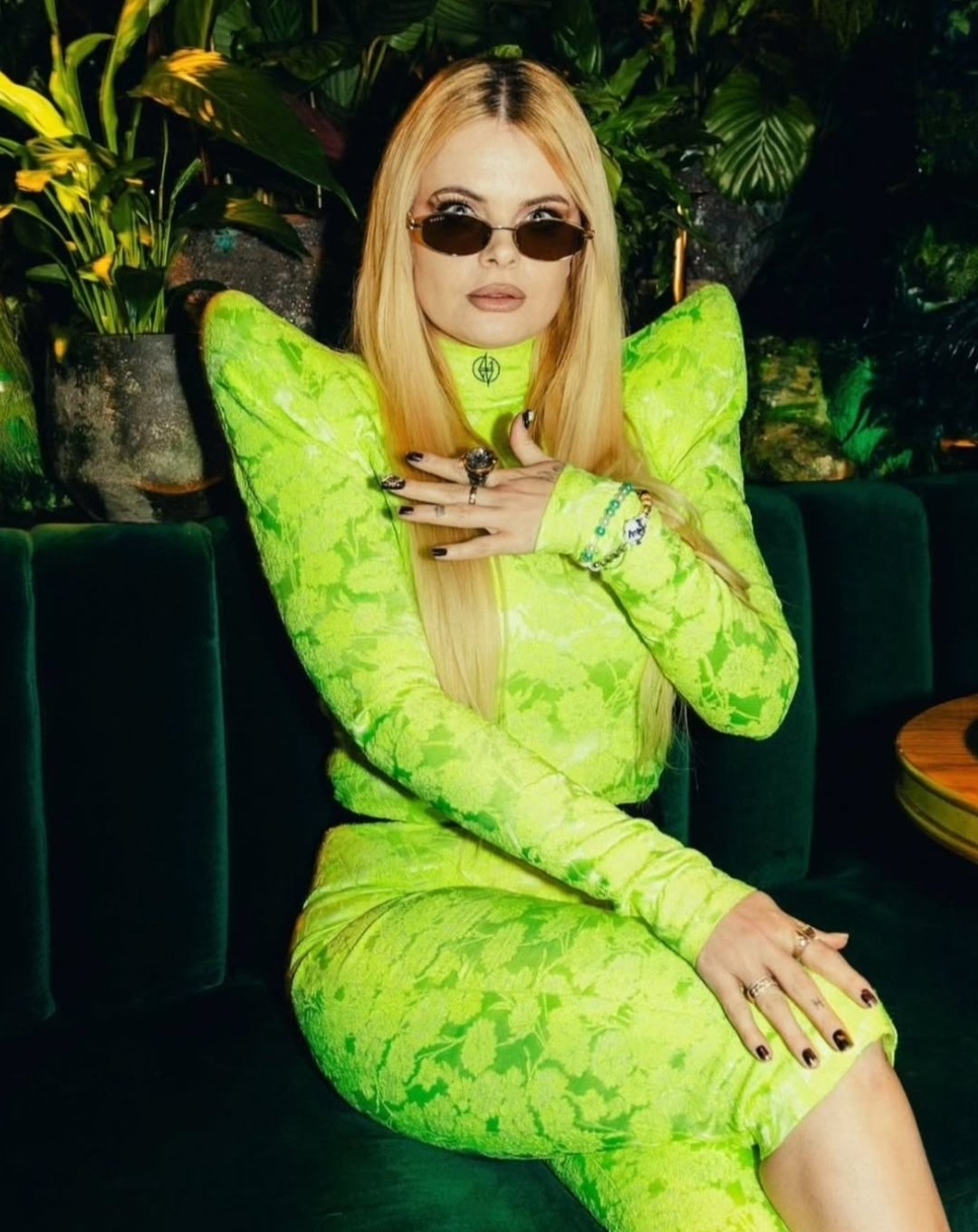 Woman with long blonde hair wearing a neon green patterned outfit, dark sunglasses, and multiple rings and bracelets, sitting on a green velvet sofa surrounded by lush green plants.