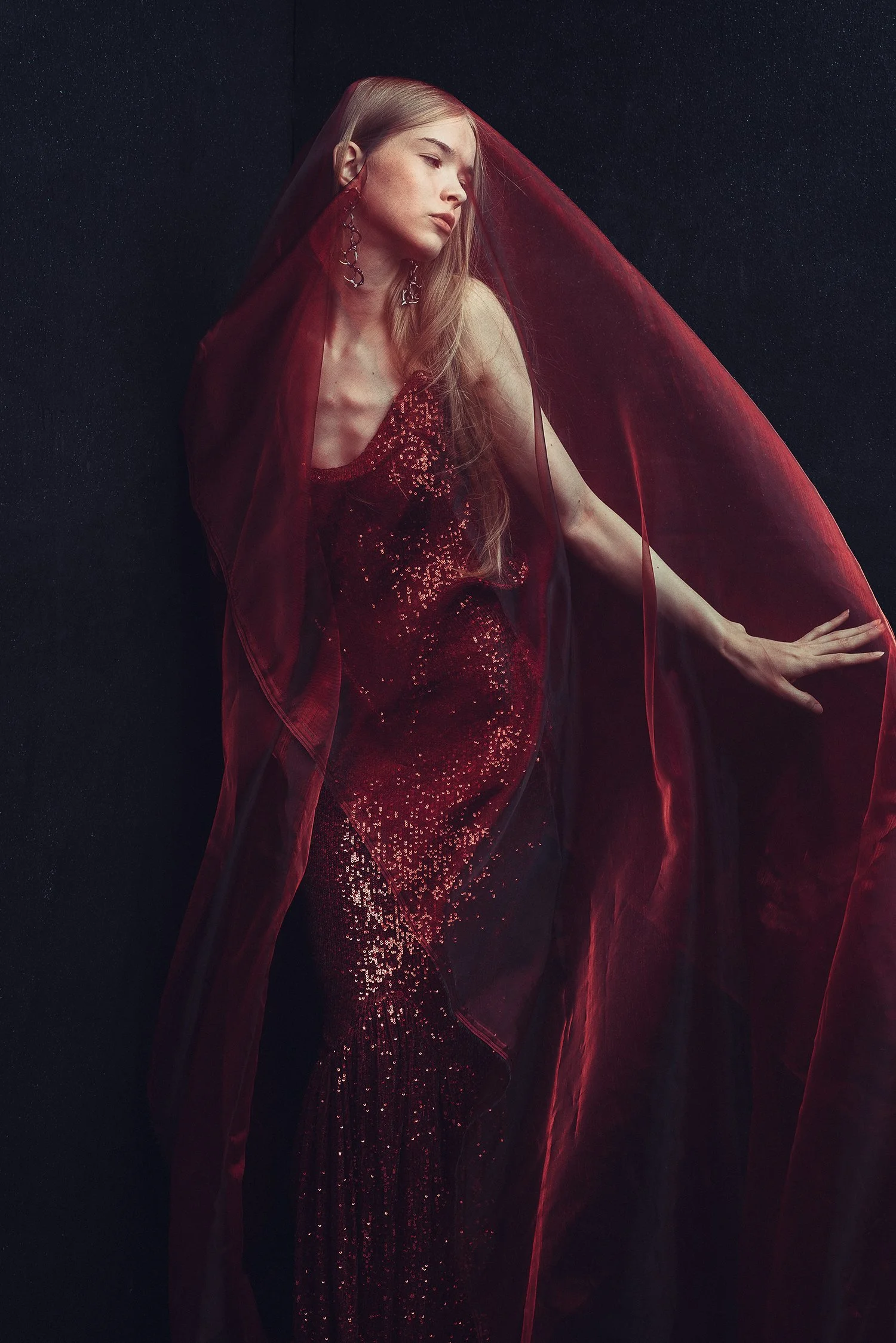 A woman in a long, shimmering red dress with a red veil draped over her head, posing against a dark background.