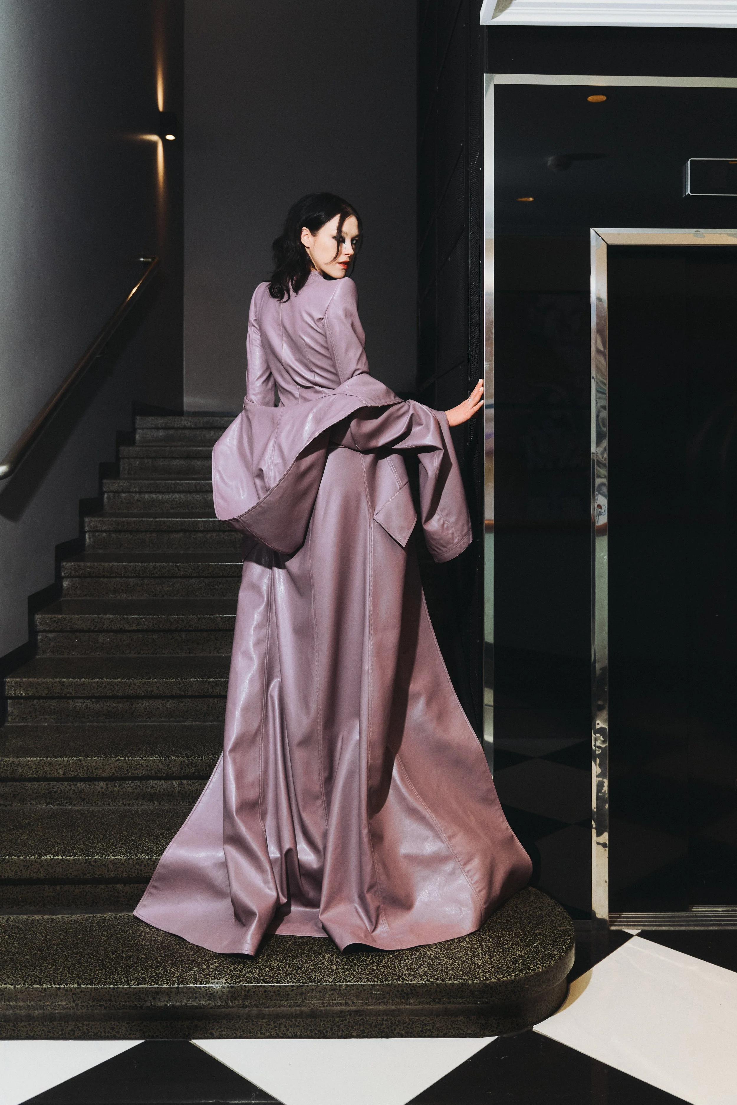A woman in a long, mauve-colored dress stands on a stairway, holding the wall with one hand and turning her head to look back.