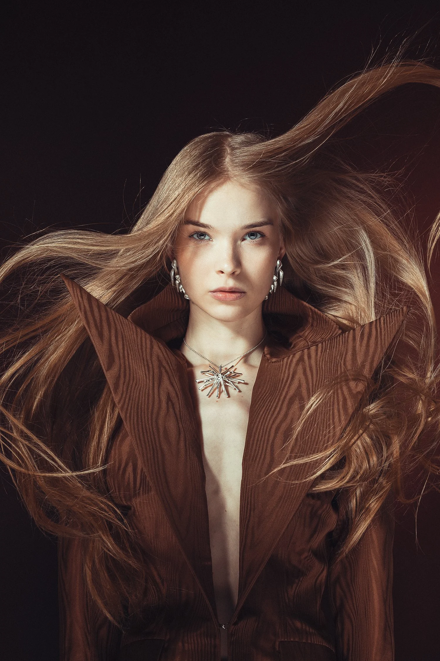 A woman with long, flowing blonde hair wearing a brown blazer with a large collar, silver jewelry, and a statement necklace against a black background.