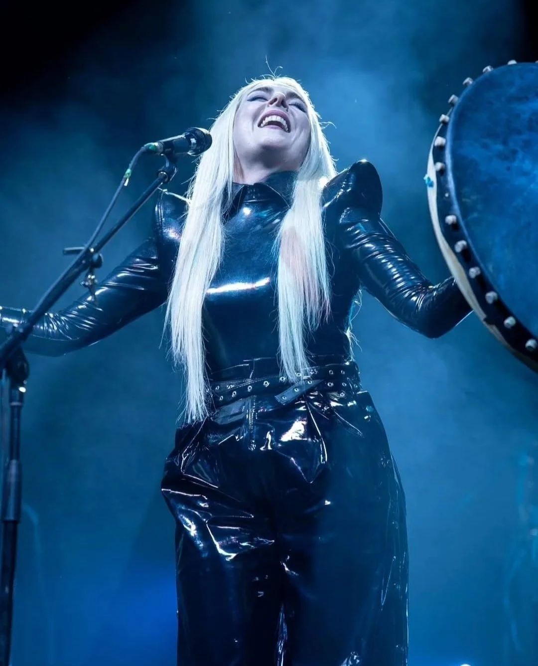 A female performer singing passionately on stage, dressed in a shiny black leather outfit, with long blonde hair and a microphone in front of her.