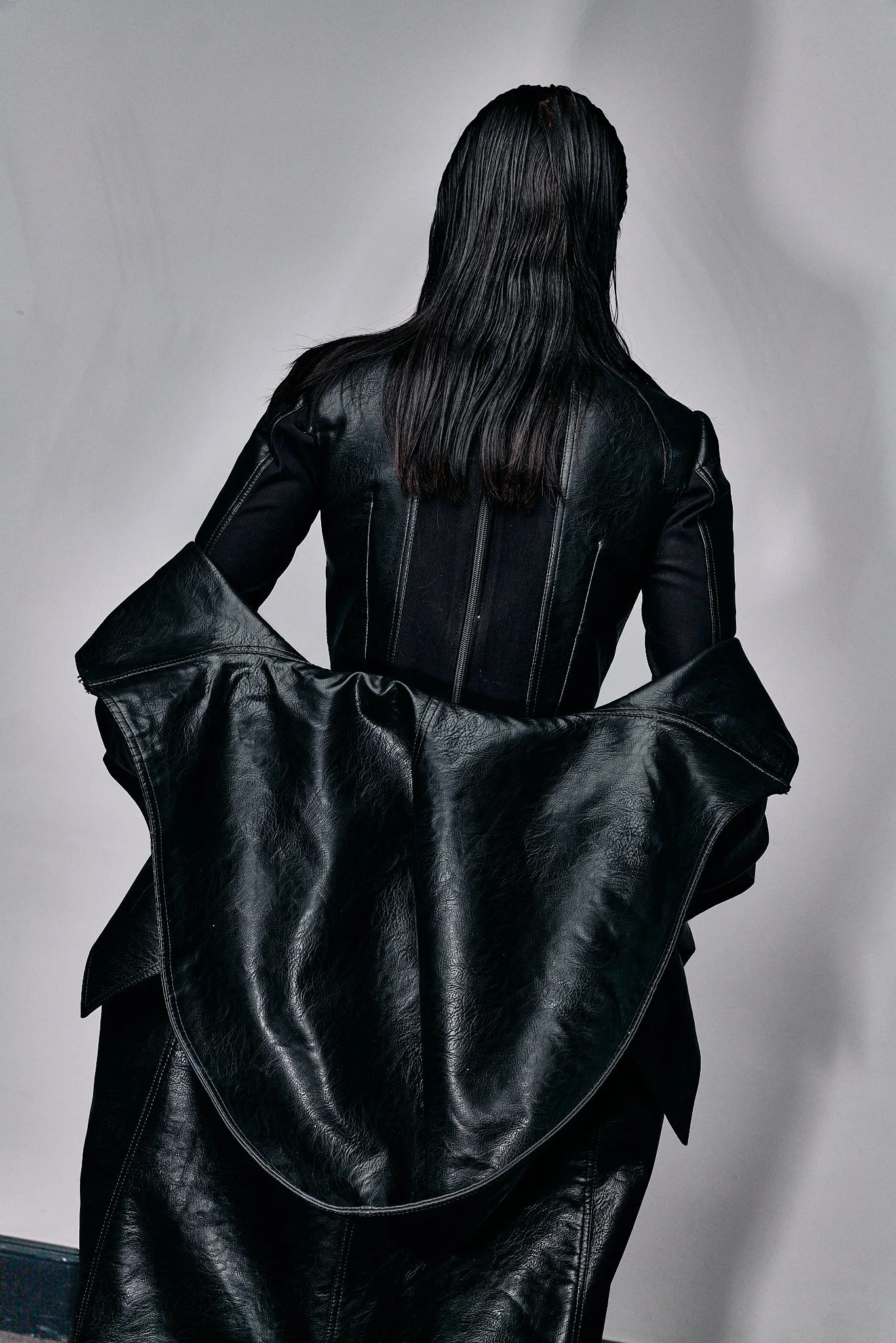 Person with long dark hair wearing a black leather jacket, holding another leather jacket in front of their lower body, standing against a plain gray background.