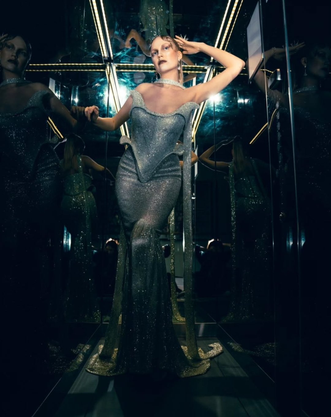 A woman in a silver, glittery gown standing in a mirrored room, surrounded by her reflections.