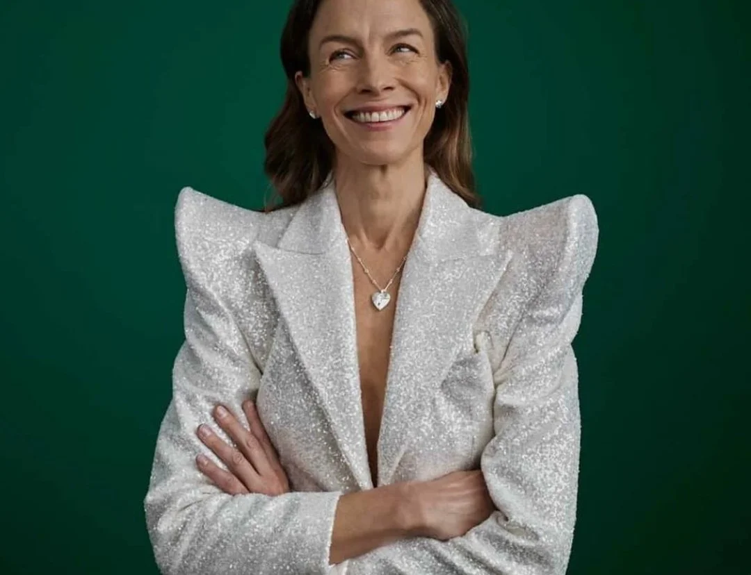 A woman smiling with her arms crossed, wearing a sparkling white blazer and a heart-shaped necklace, standing against a green background.