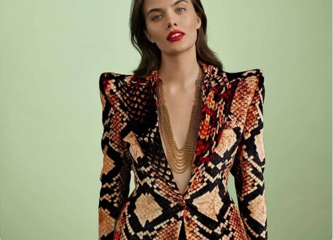 A woman with dark hair wearing a colorful snake-print blazer with shoulder pads, deep V-neck, and layered necklaces, standing against a green background.