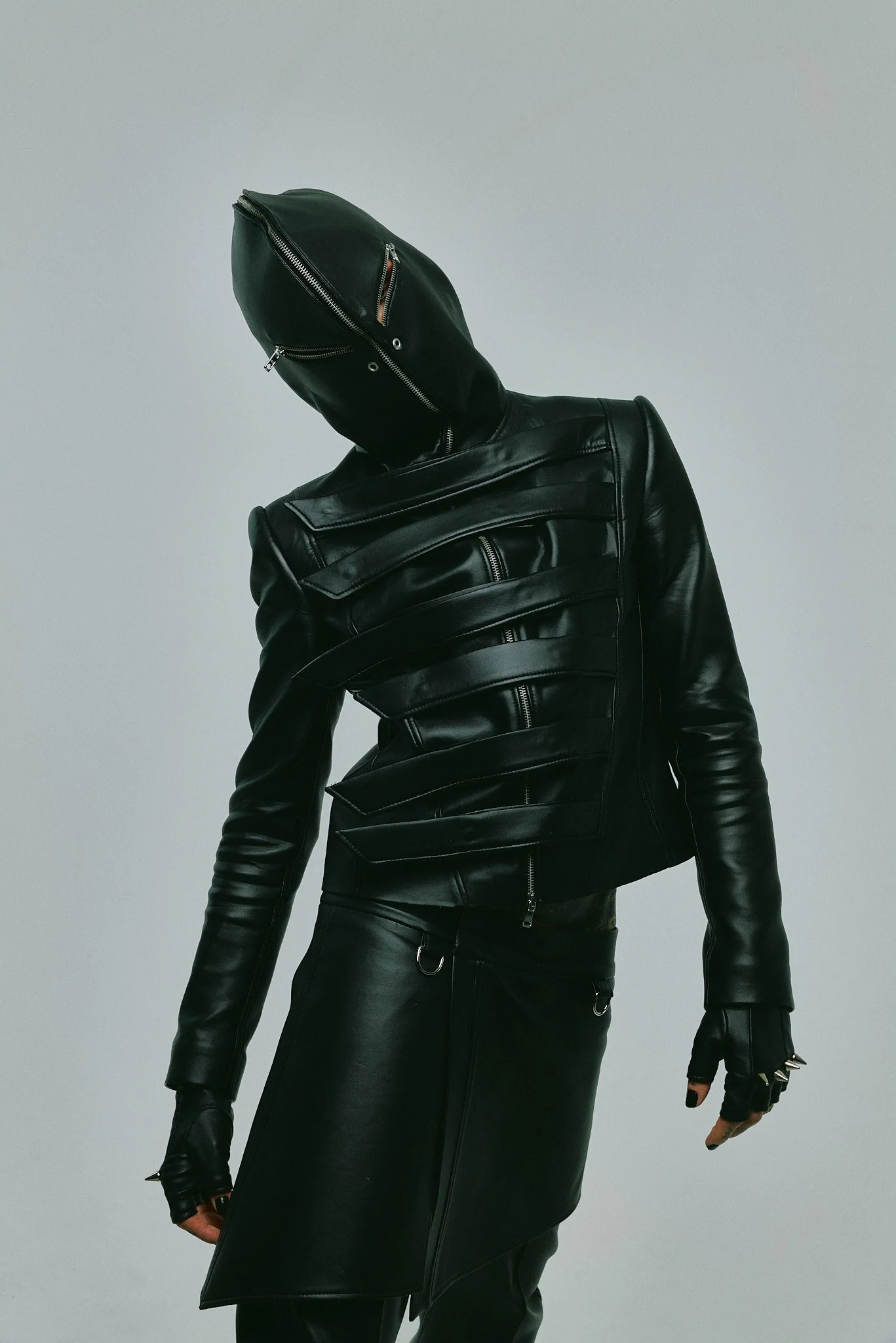 Person wearing black leather jacket, black leather skirt, black gloves, and a black face covering with zippers, standing against a plain light background.