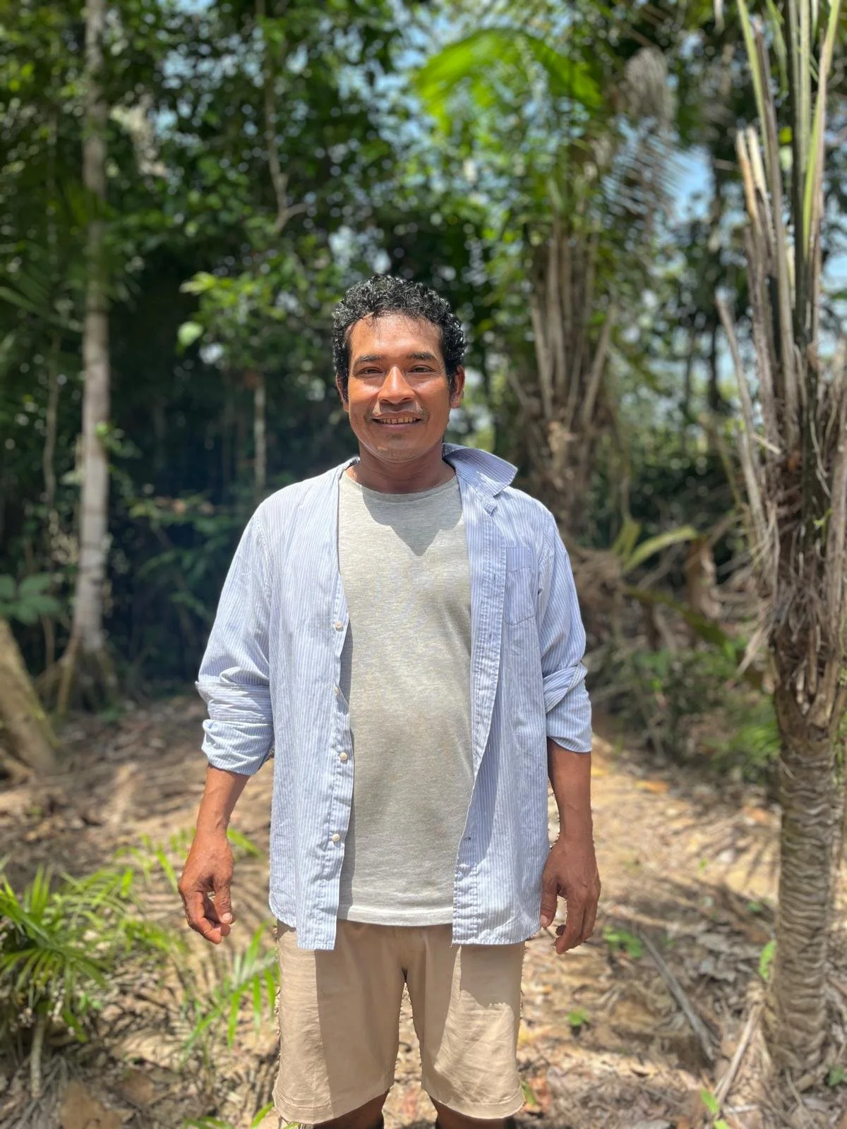 Man standing in a lush green jungle, smiling, wearing a light blue striped shirt over a gray t-shirt and beige shorts.