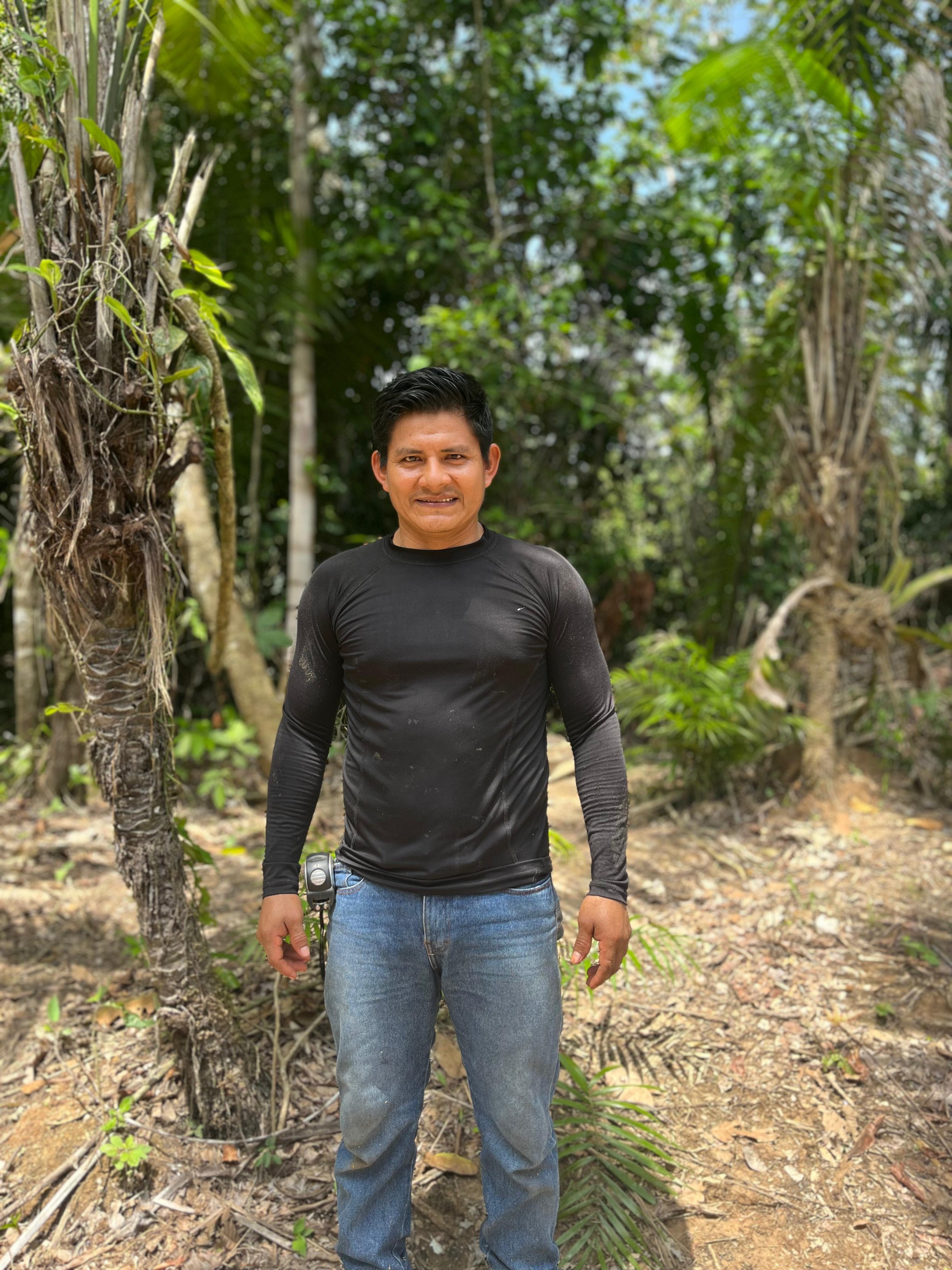 A man with black hair wearing a black long-sleeve shirt and blue jeans standing outdoors in a forested area with green trees and plants.