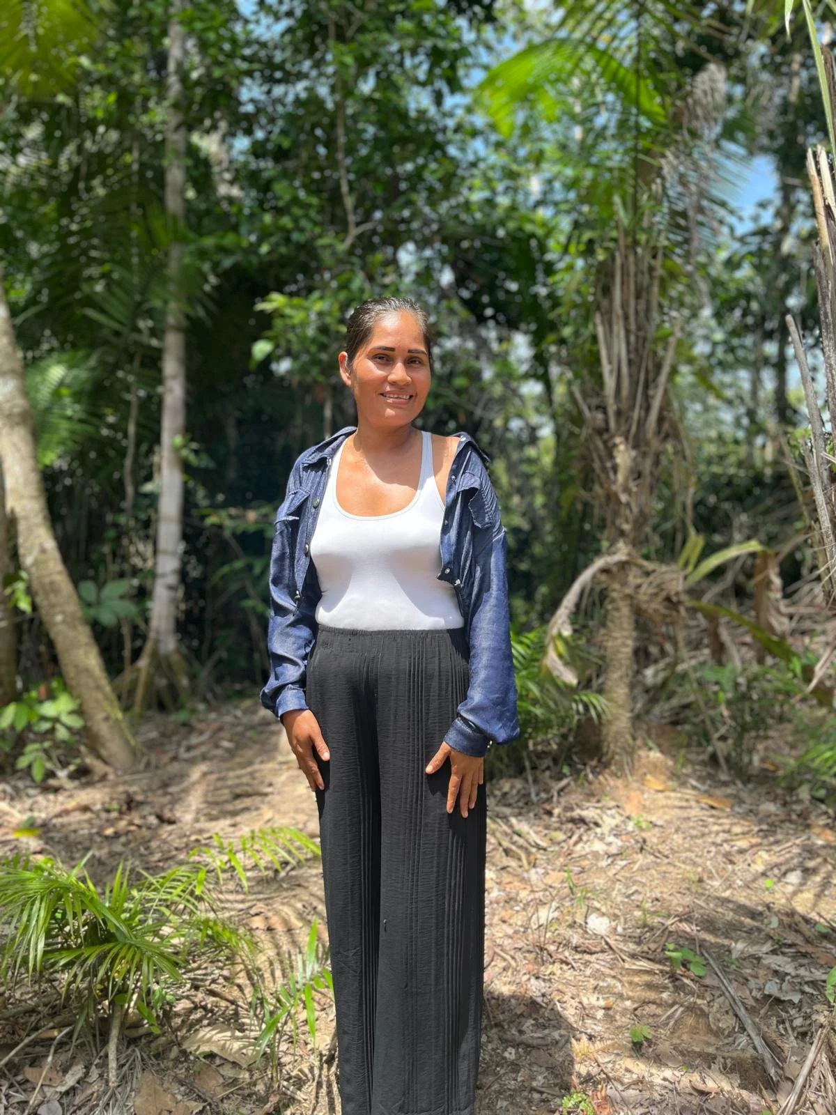 A woman standing on a dirt path in a lush tropical forest, smiling, wearing a white tank top, black wide-leg pants, and a blue denim jacket.