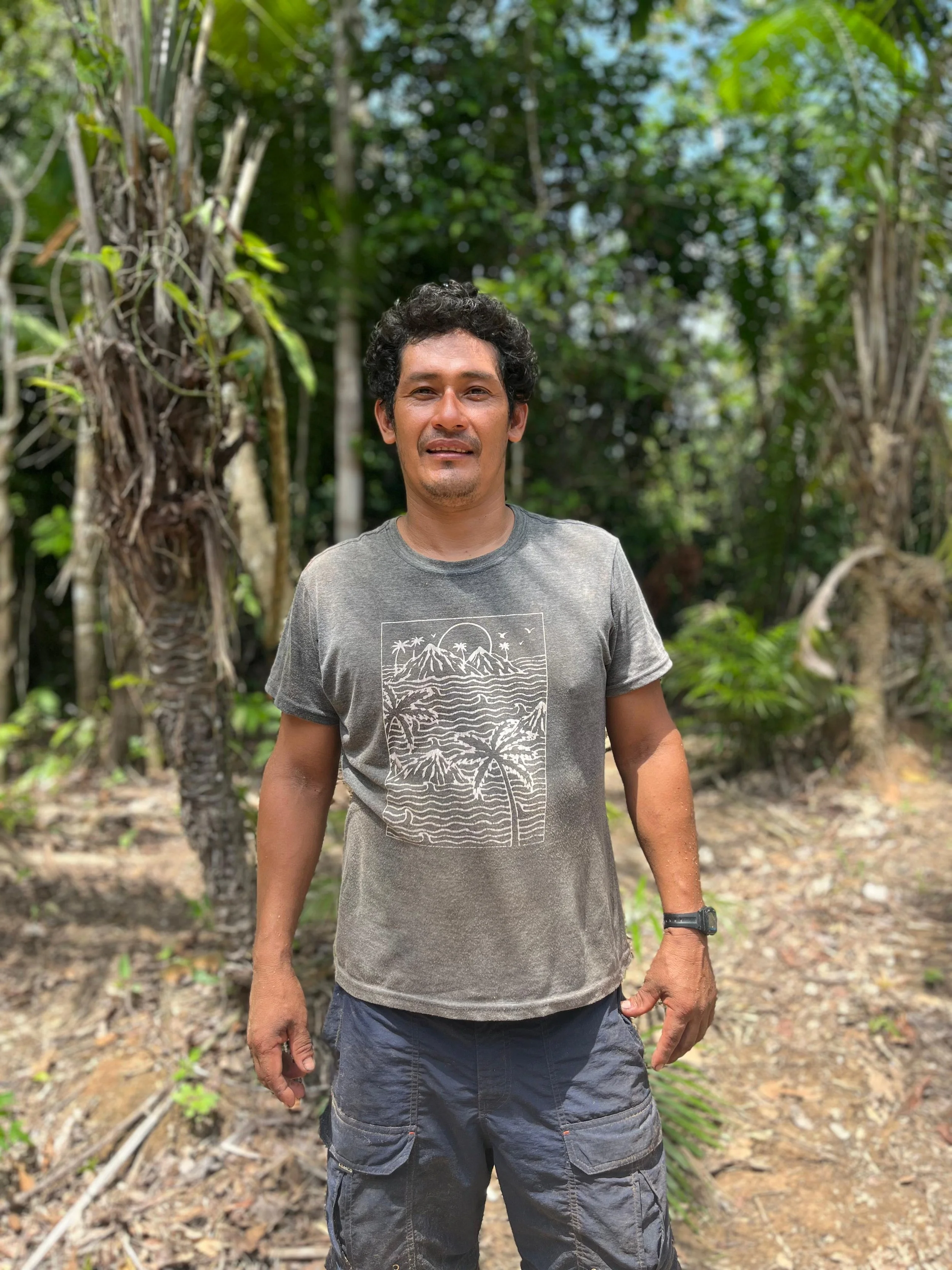 A man standing outdoors in a jungle or forest setting, wearing a gray t-shirt with a mountain and palm tree design, and dark cargo shorts.