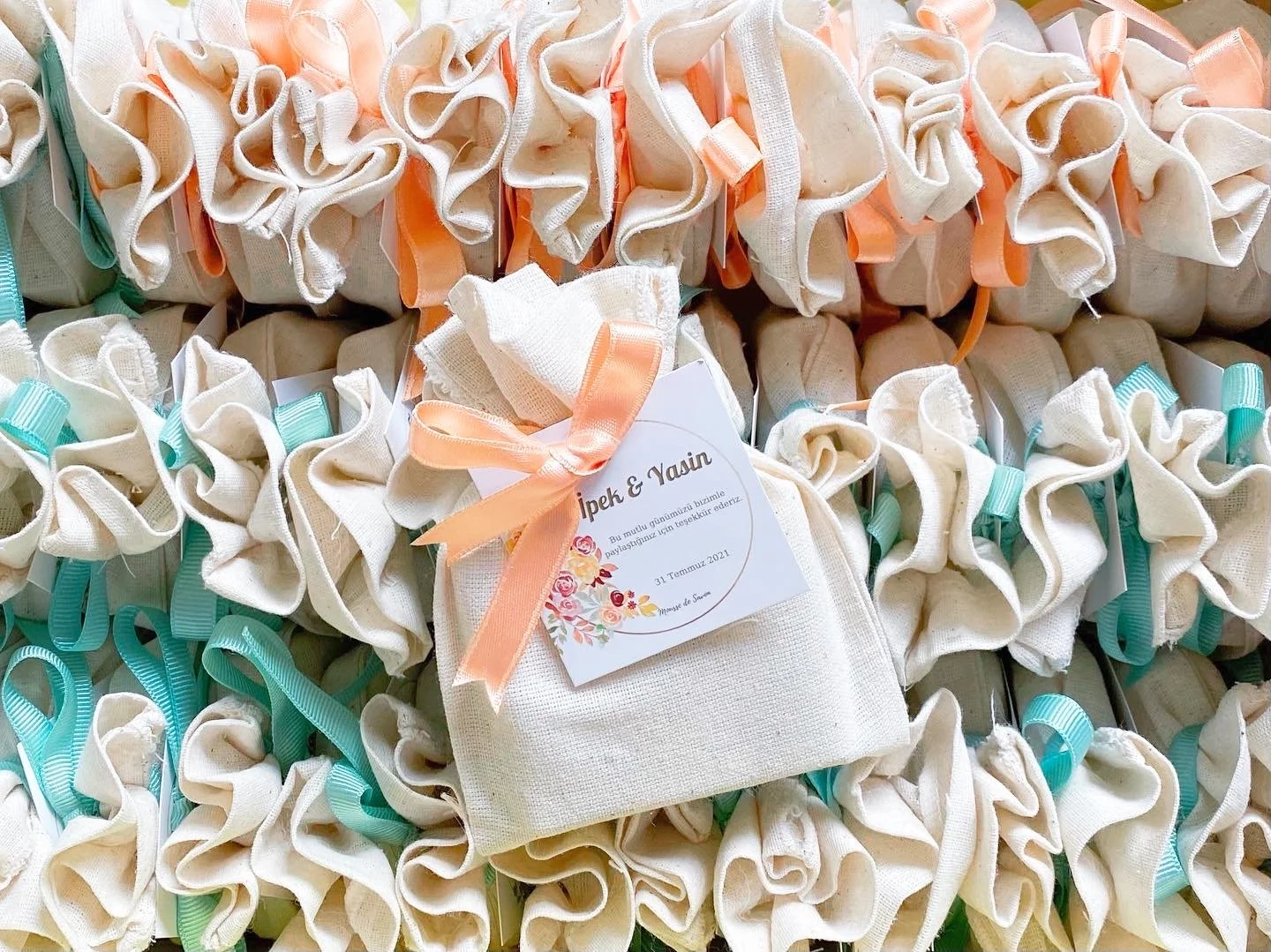 A collection of beige fabric pouches with colorful ribbons tied around them, featuring a small card with Turkish text and floral decoration.