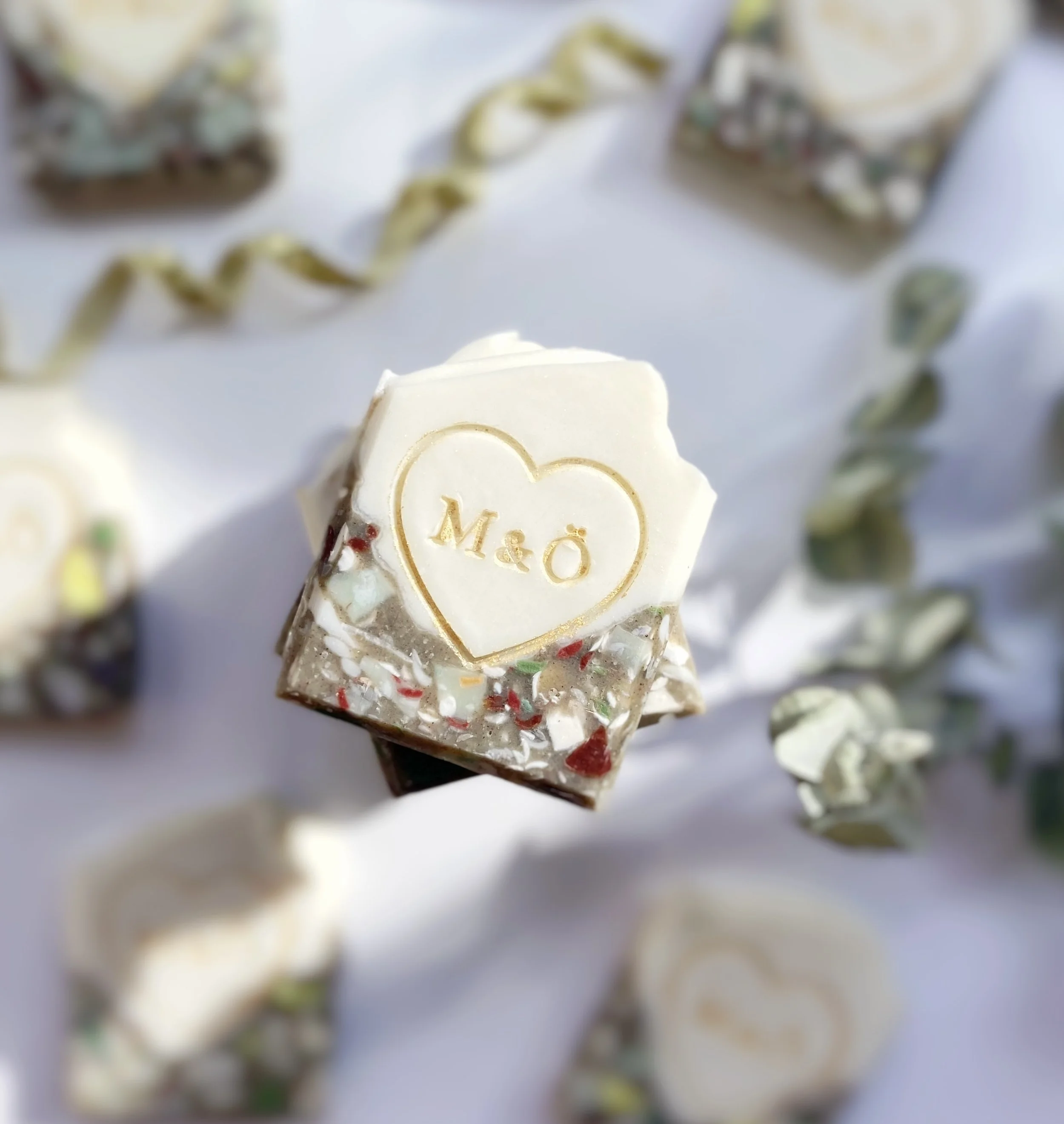 A decorated chocolate piece topped with white chocolate, inscribed with a heart and the words 'M & Ö', surrounded by similar chocolates and a white ribbon on a white surface.