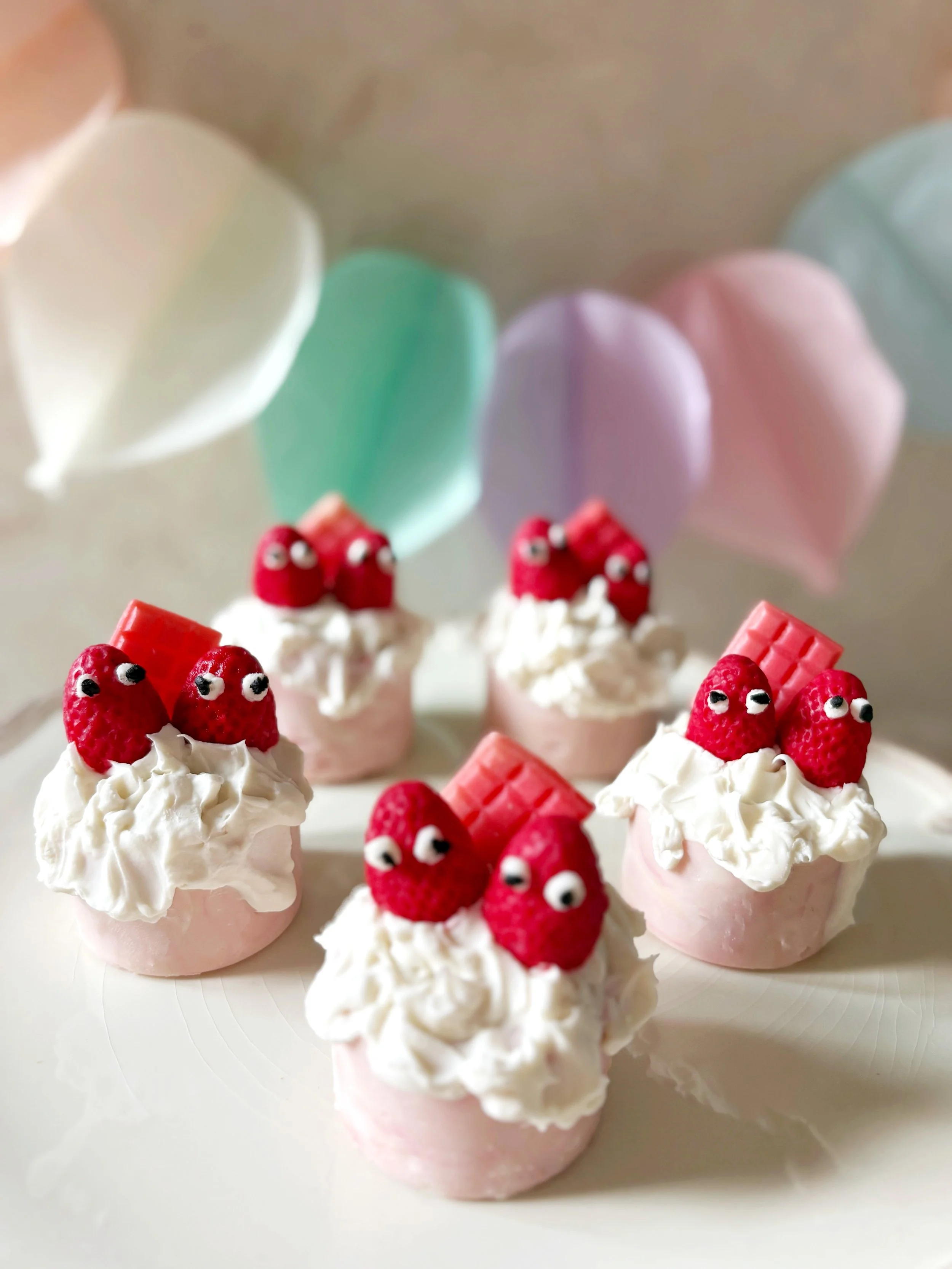 Mini pink cupcakes decorated with whipped cream, strawberries with googly eyes, and pink chocolate bars, with pastel balloons in the background.