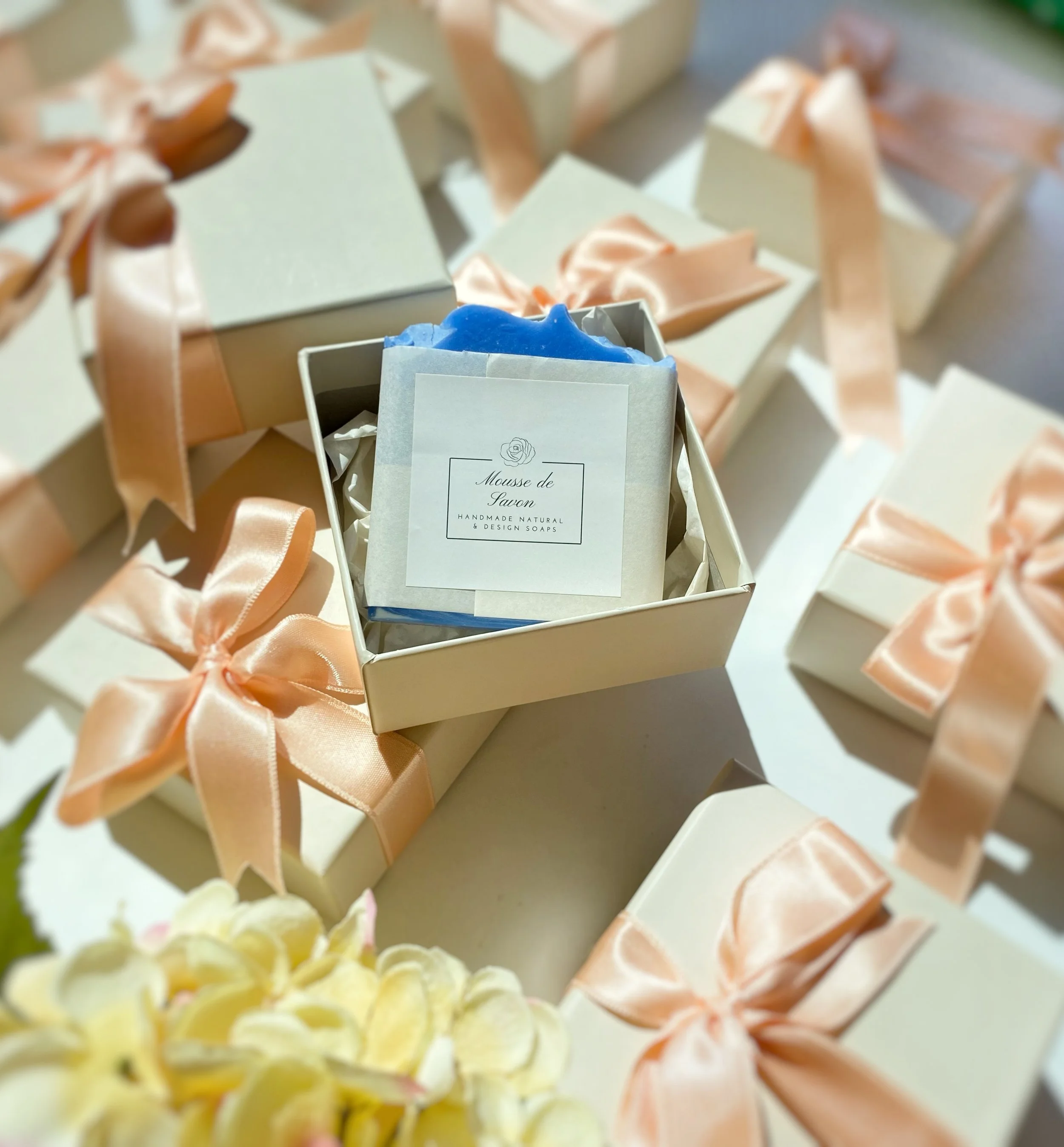 Arranged gift boxes decorated with peach satin ribbons, with one box open revealing blue handmade soap labeled "Mousse de Savon".