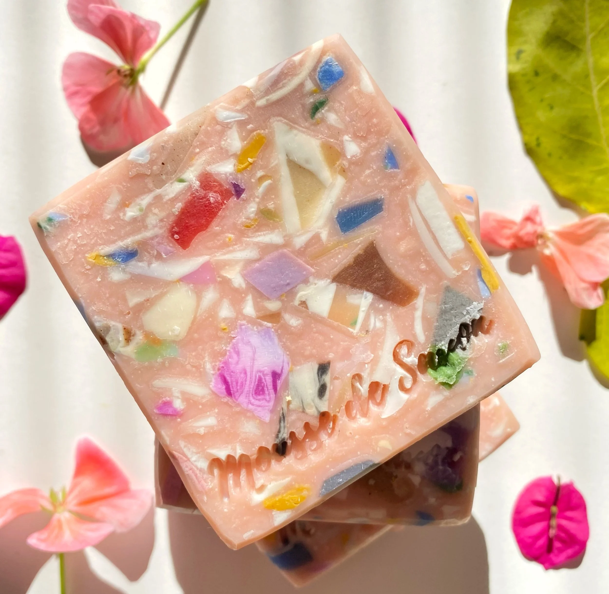 Close-up of a pink soap bar with colorful embedded shapes and the word 'mama soap' engraved on it, surrounded by pink, purple, and green flowers.