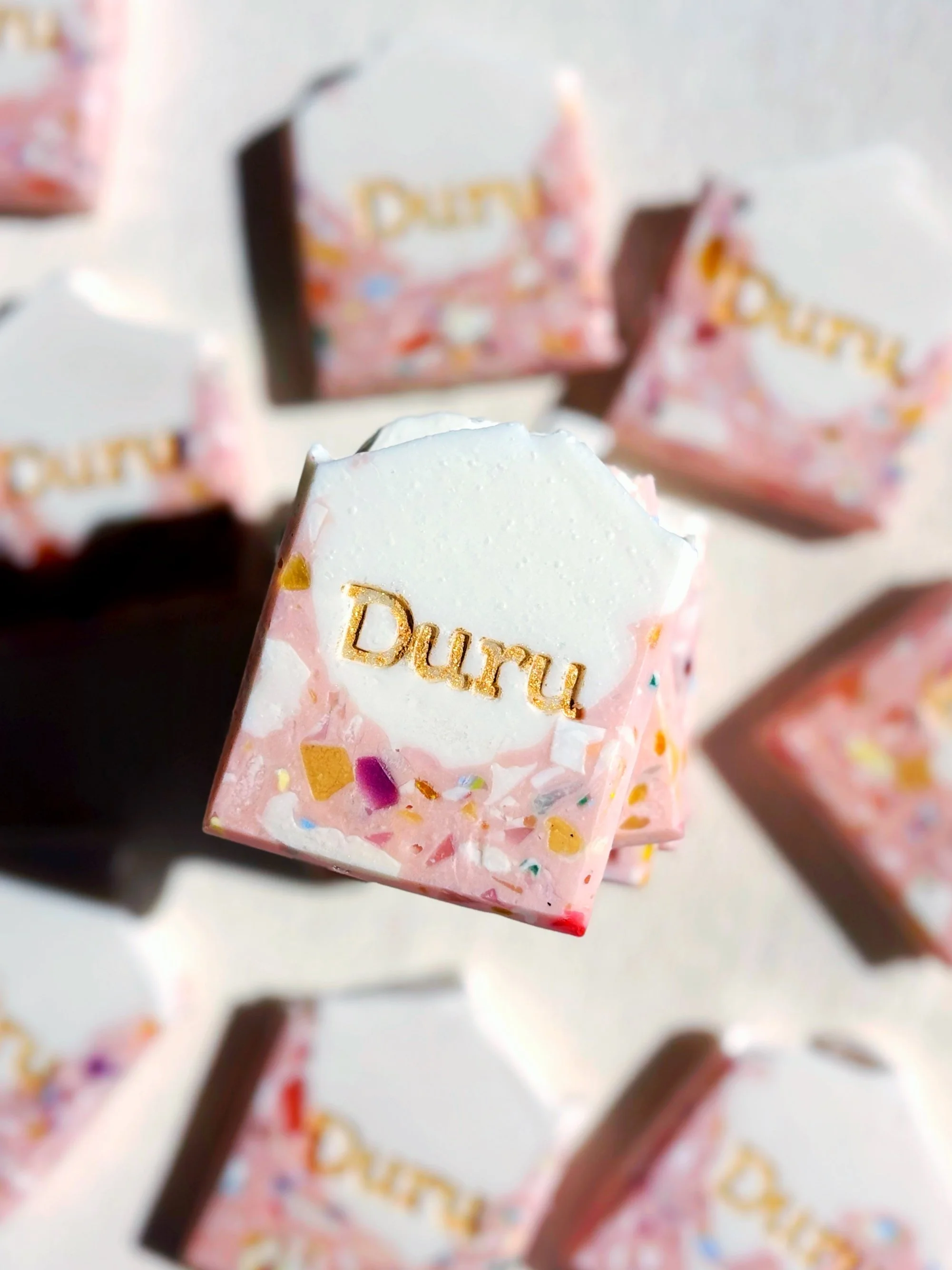 Close-up of a pink and white praline with the word 'Duru' embossed in gold, surrounded by other chocolates with similar branding.