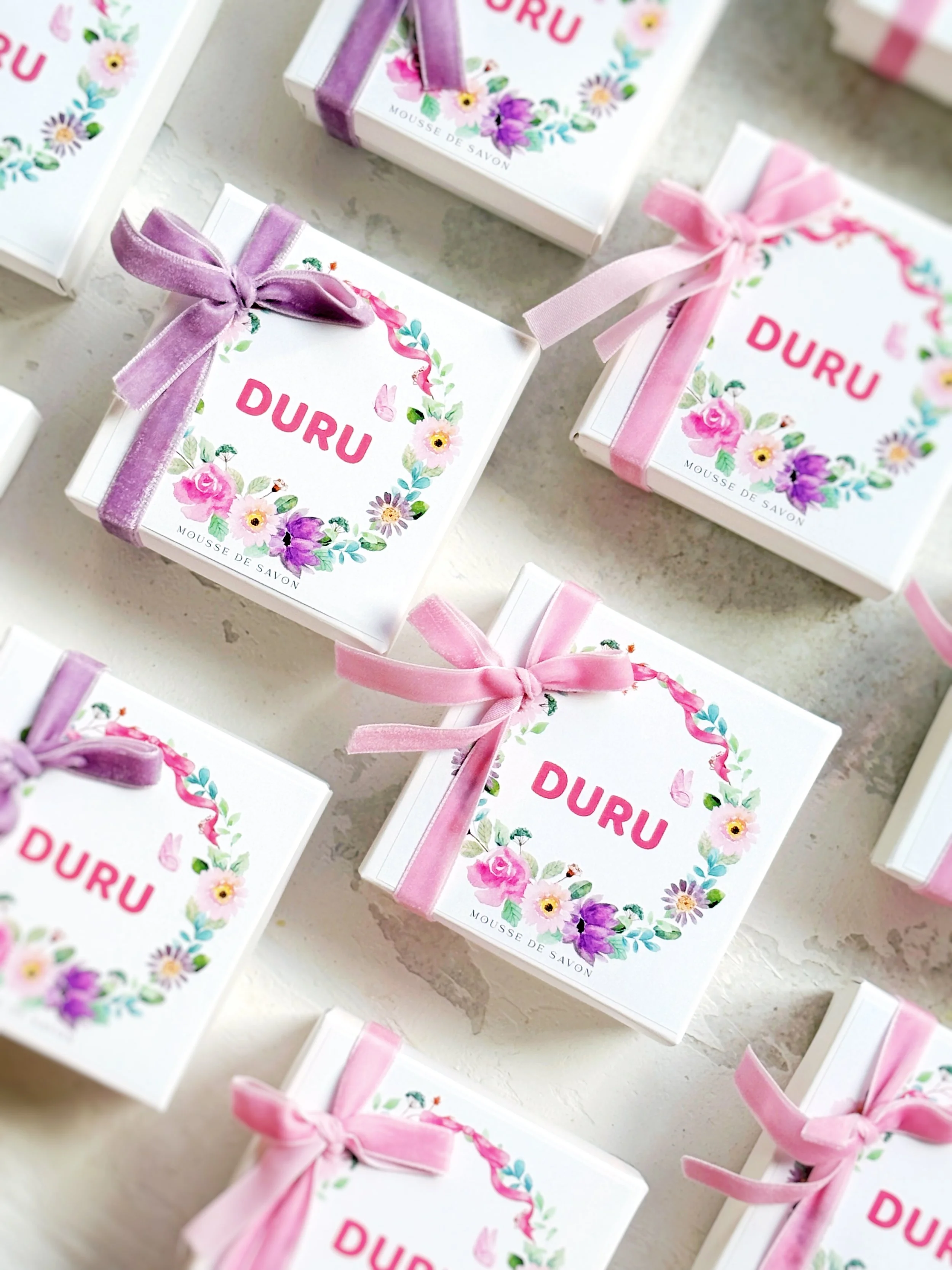 Boxes of soap labeled "DURU" with pink and purple ribbons, decorated with floral wreaths and small butterflies on a light surface.