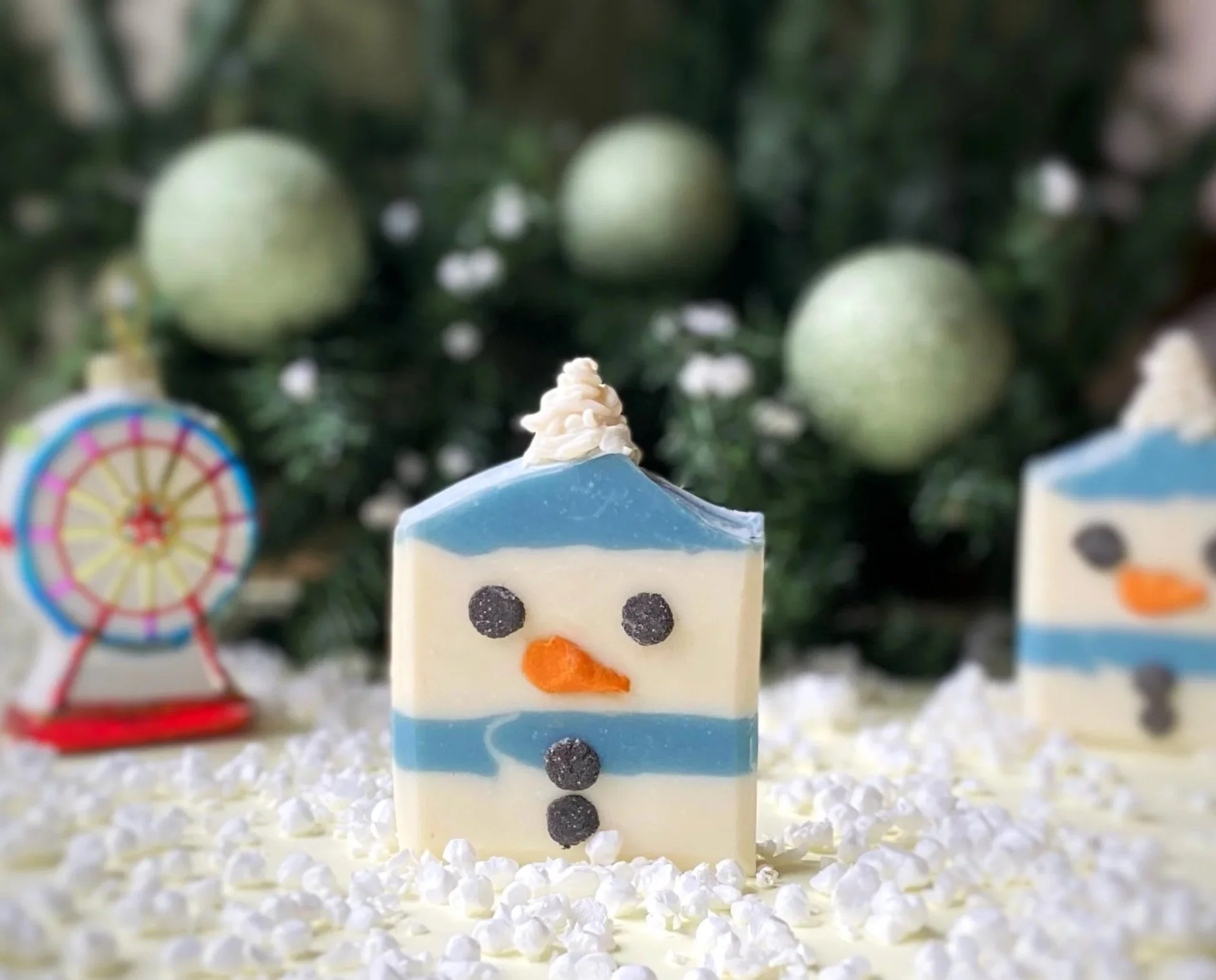 Decorative snowman-shaped confection with a white face, black eyes, an orange nose, and a blue and white hat, surrounded by festive holiday decorations.