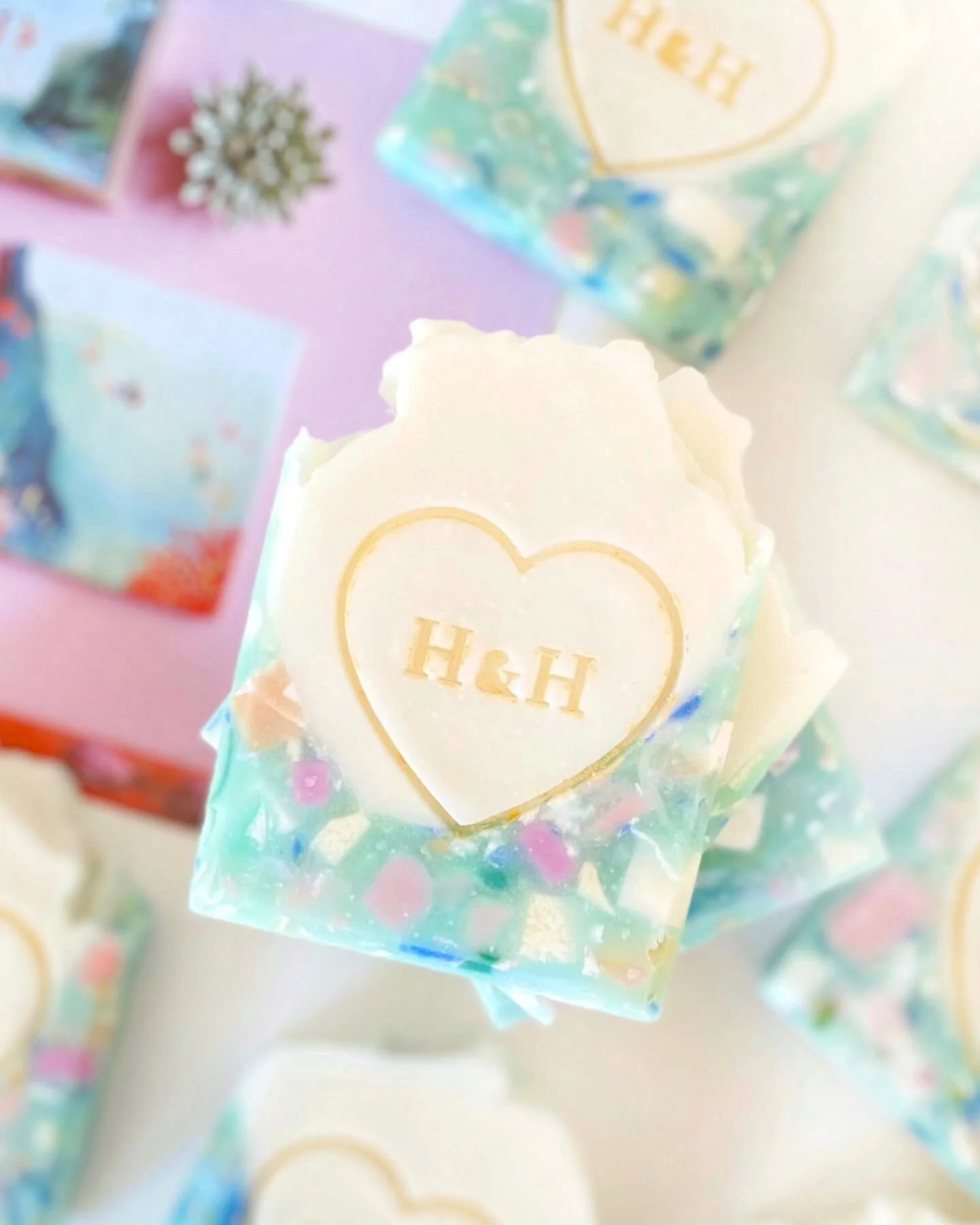 Colorful soap with a white heart and the initials H&H engraved inside. The soap contains small, colorful decorative bits. Background shows packaging and other soap bars with matching themes.