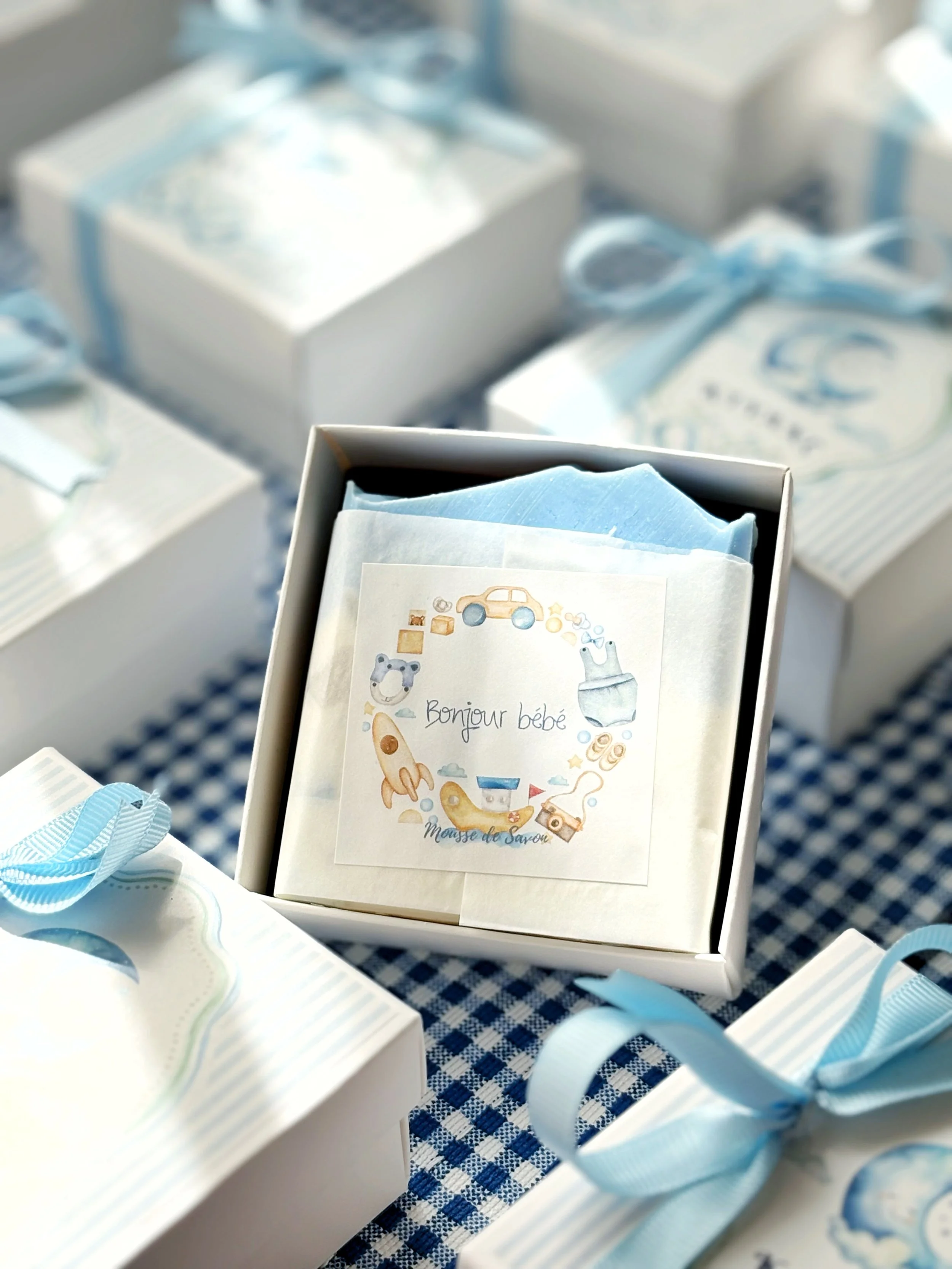 Baby shower favor box with a label that reads 'Bonjour bébé' and illustrations of a bear, rocket, camera, and toys on a blue and white color theme.