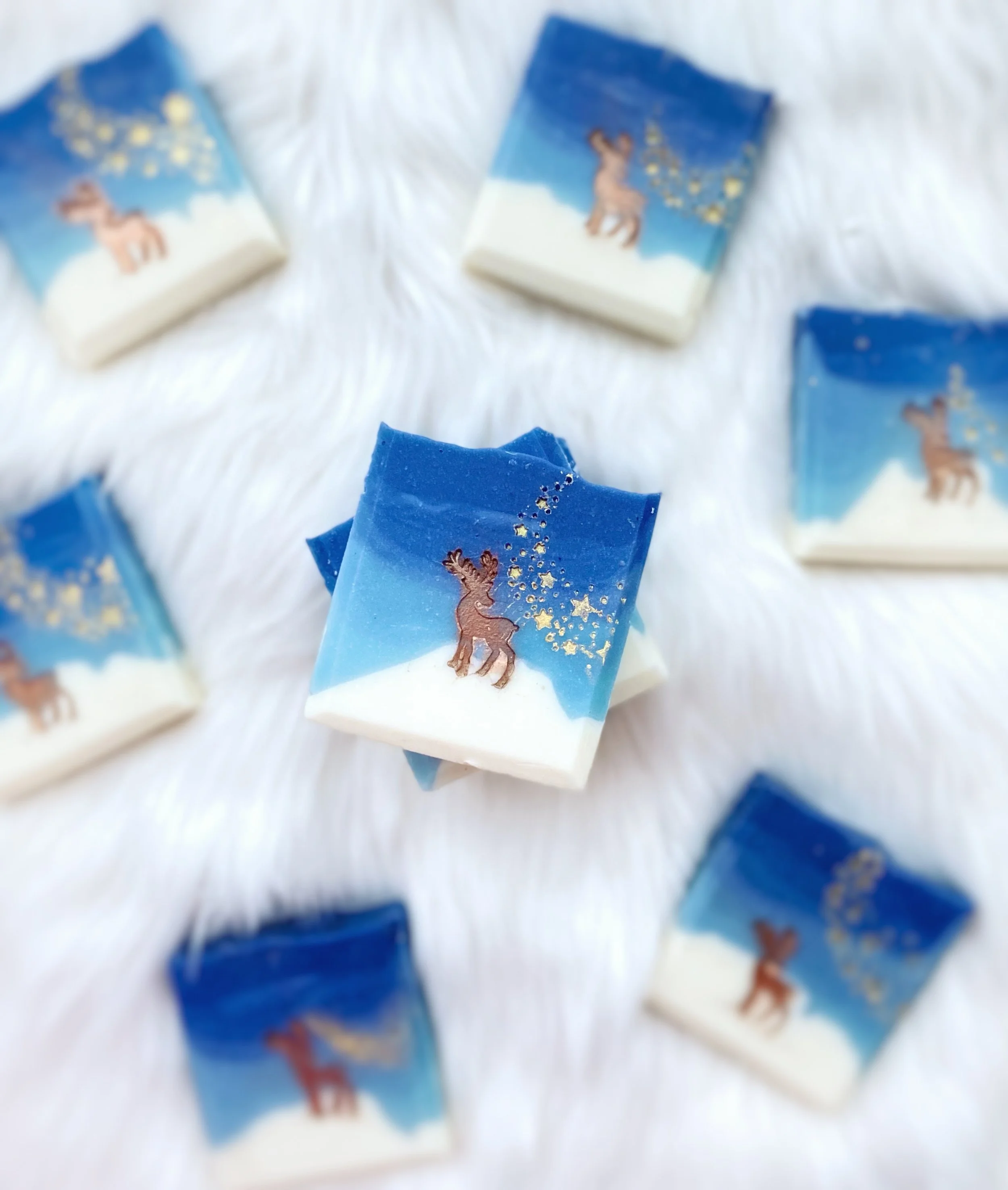 Christmas-themed soap bars with a blue and white gradient background, decorated with a reindeer and gold stars, arranged on a white fluffy surface.