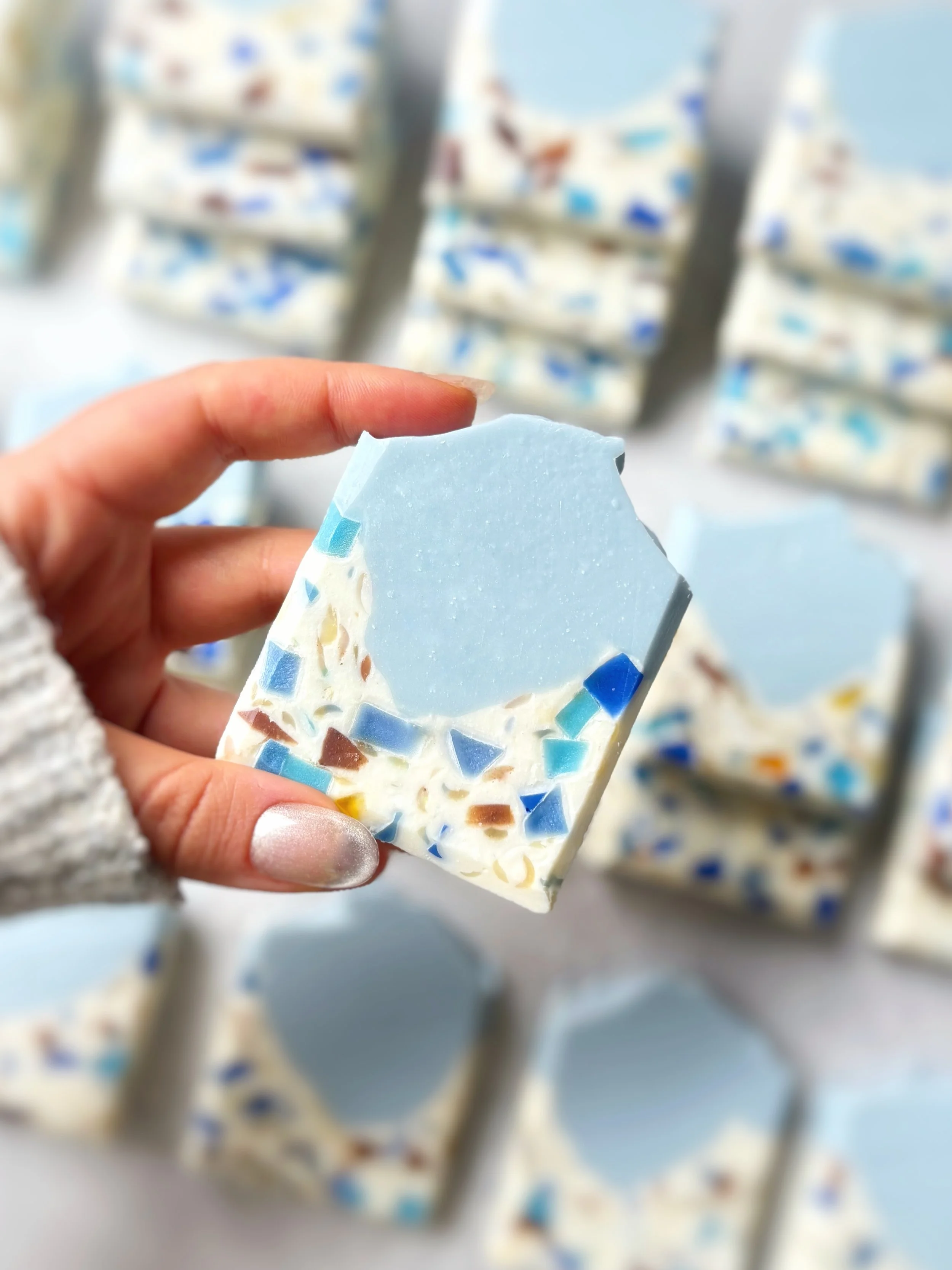 Hand holding a bar of soap with blue, white, and colored mosaic-like design, blurred background of similar soap bars.