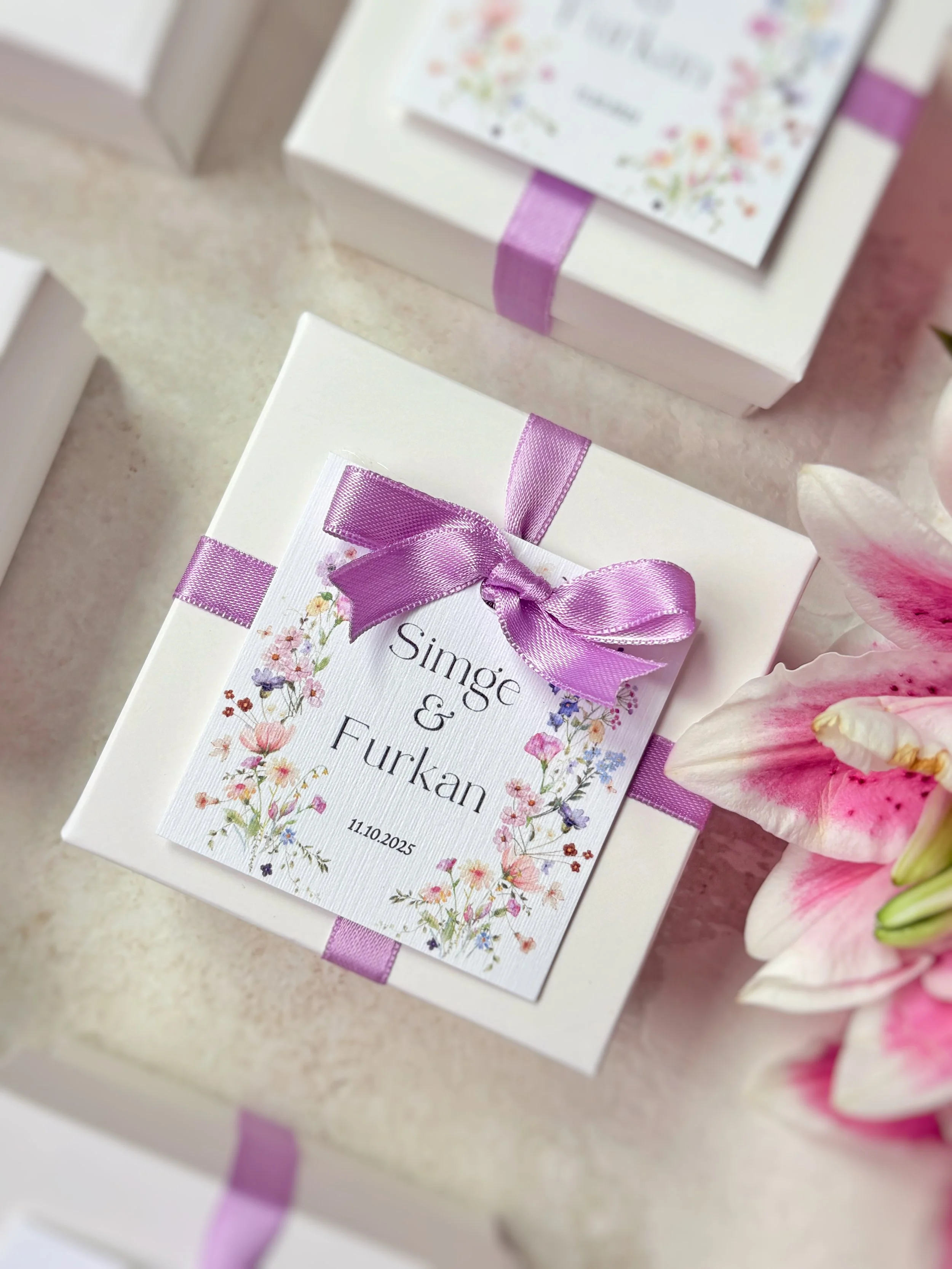 White gift box decorated with lavender ribbon and a floral event label reading 'Singe & Furkan 11.10.2025' with pink lilies beside it.