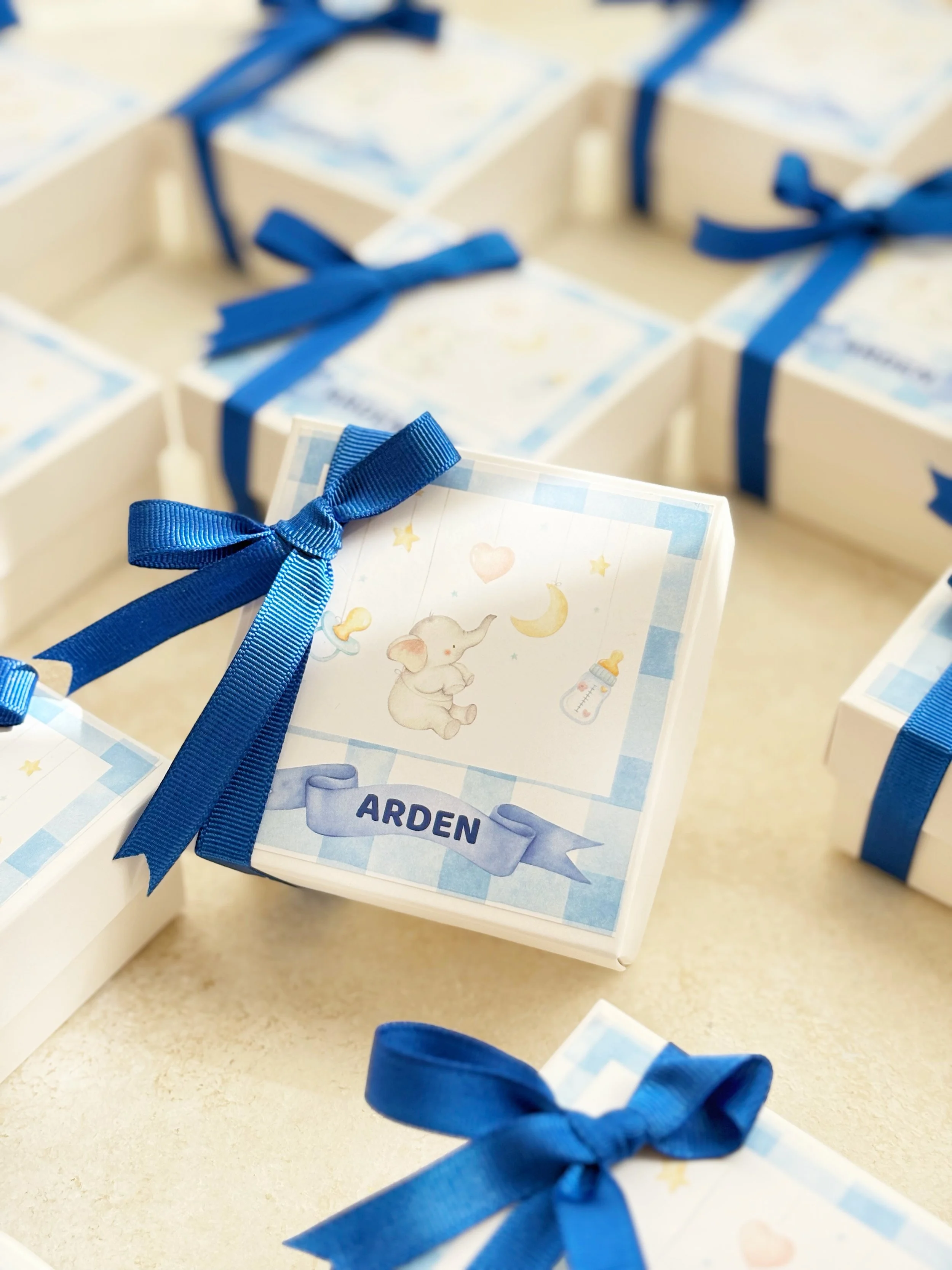 Baby shower favor boxes decorated with blue ribbons, featuring a label with a cute elephant illustration, a yellow heart, moon, and baby bottle, and the name 'Arden'.