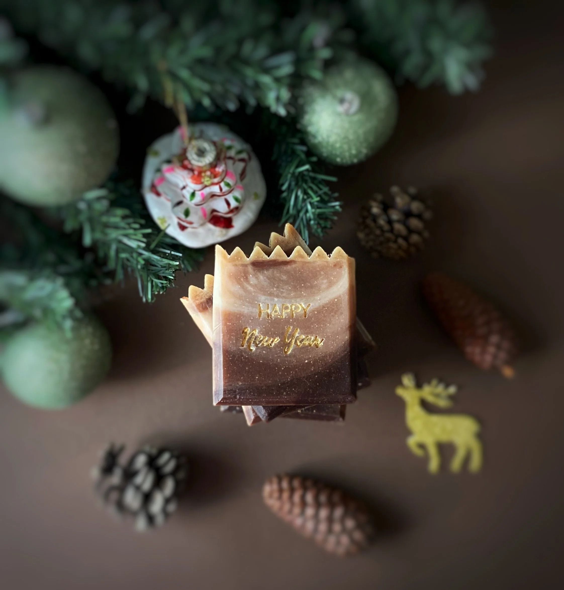 A chocolate bar with the words 'HAPPY New Year' engraved on it, placed among pine cones, a small yellow reindeer cutout, and Christmas ornaments on a brown surface, with a decorated Christmas tree in the background.