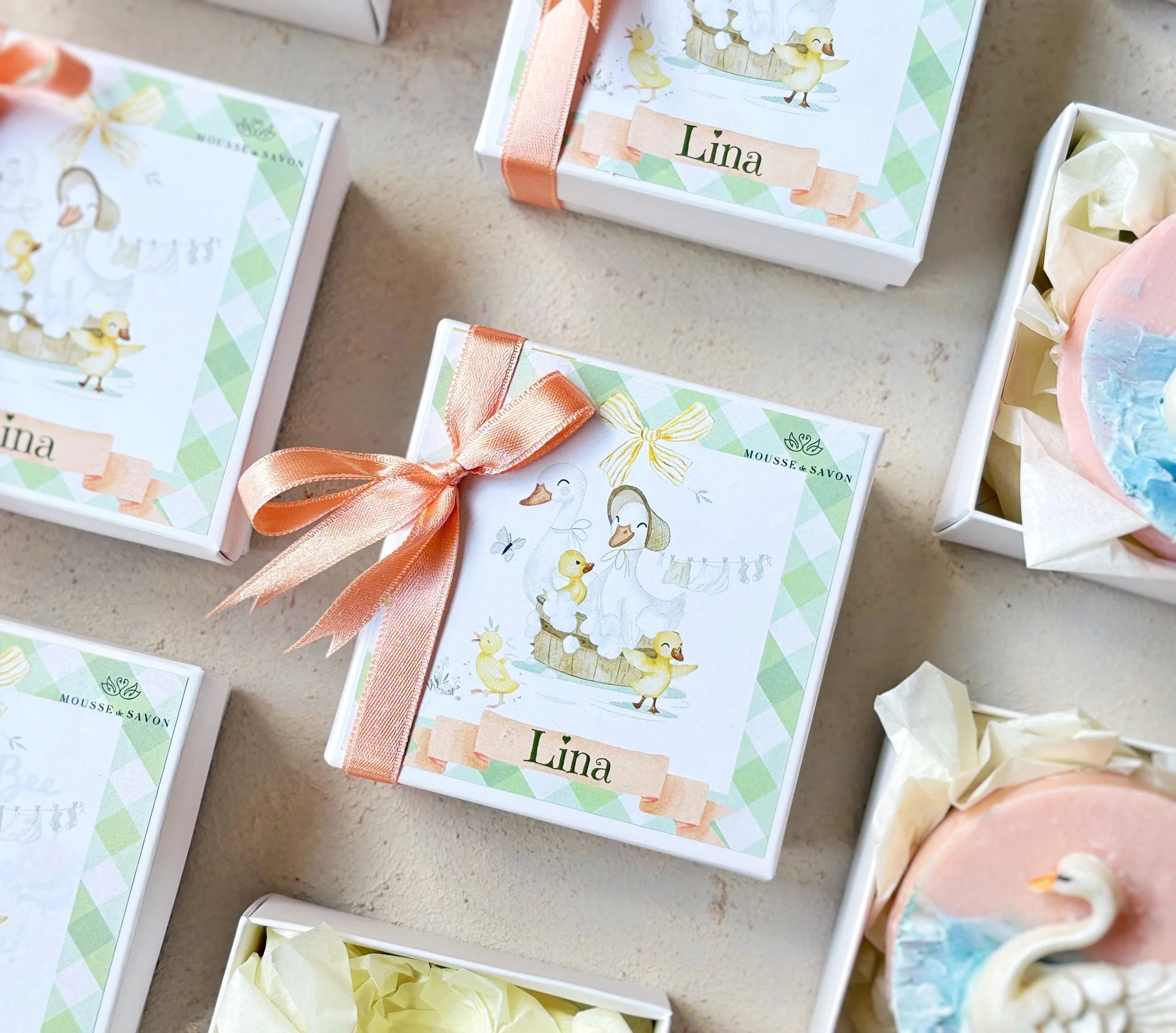 Boxed soap labeled "Lina" with a duck and goose design, tied with a peach ribbon, surrounded by other soap boxes and a swan-shaped soap in a display.